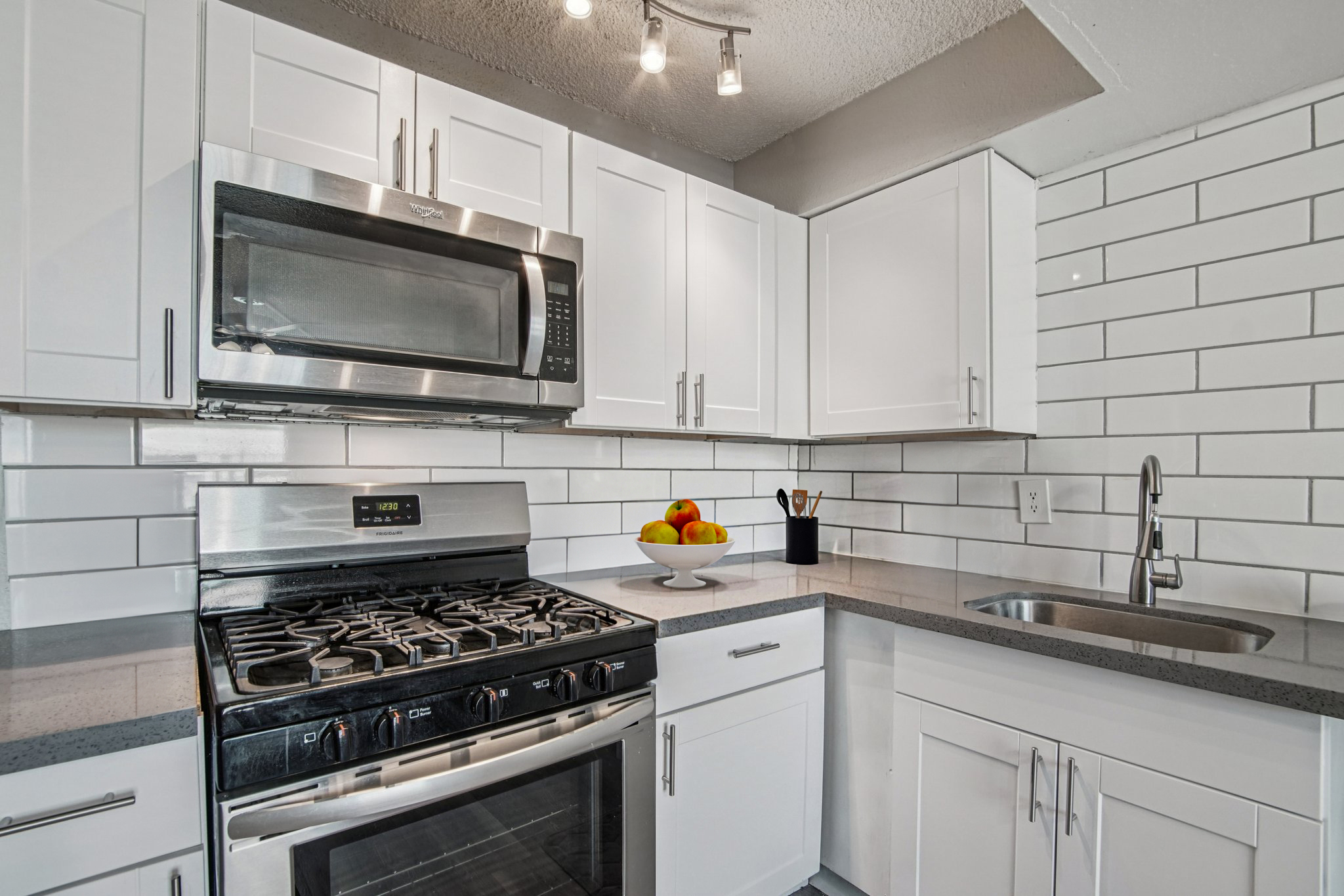 A modern kitchen featuring white cabinetry, stainless steel appliances, and a gray countertop. A microwave is mounted above a gas stove, and a bowl of colorful fruit sits on the counter. The backsplash consists of white subway tiles, and there's a sink next to the stove. Bright overhead lighting illuminates the space.