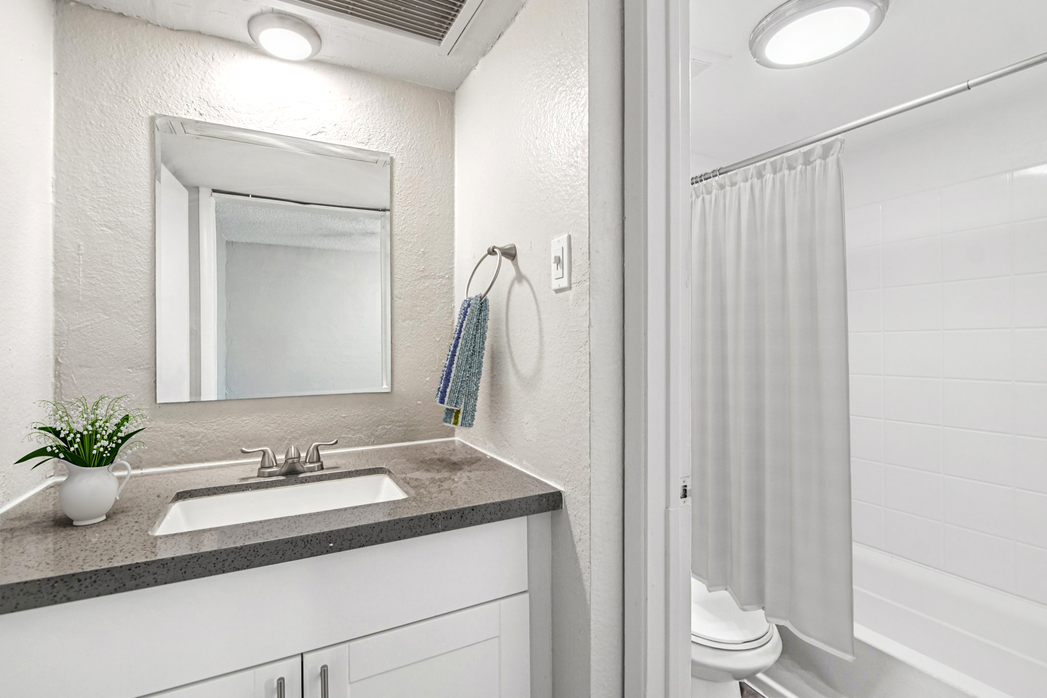 A modern bathroom featuring a sink with a gray countertop, a mirror above the sink, and a towel hanging on the wall. A potted plant sits on the countertop. The bathroom includes a shower area with a white curtain and a toilet visible in the background. The walls are painted in a light color.