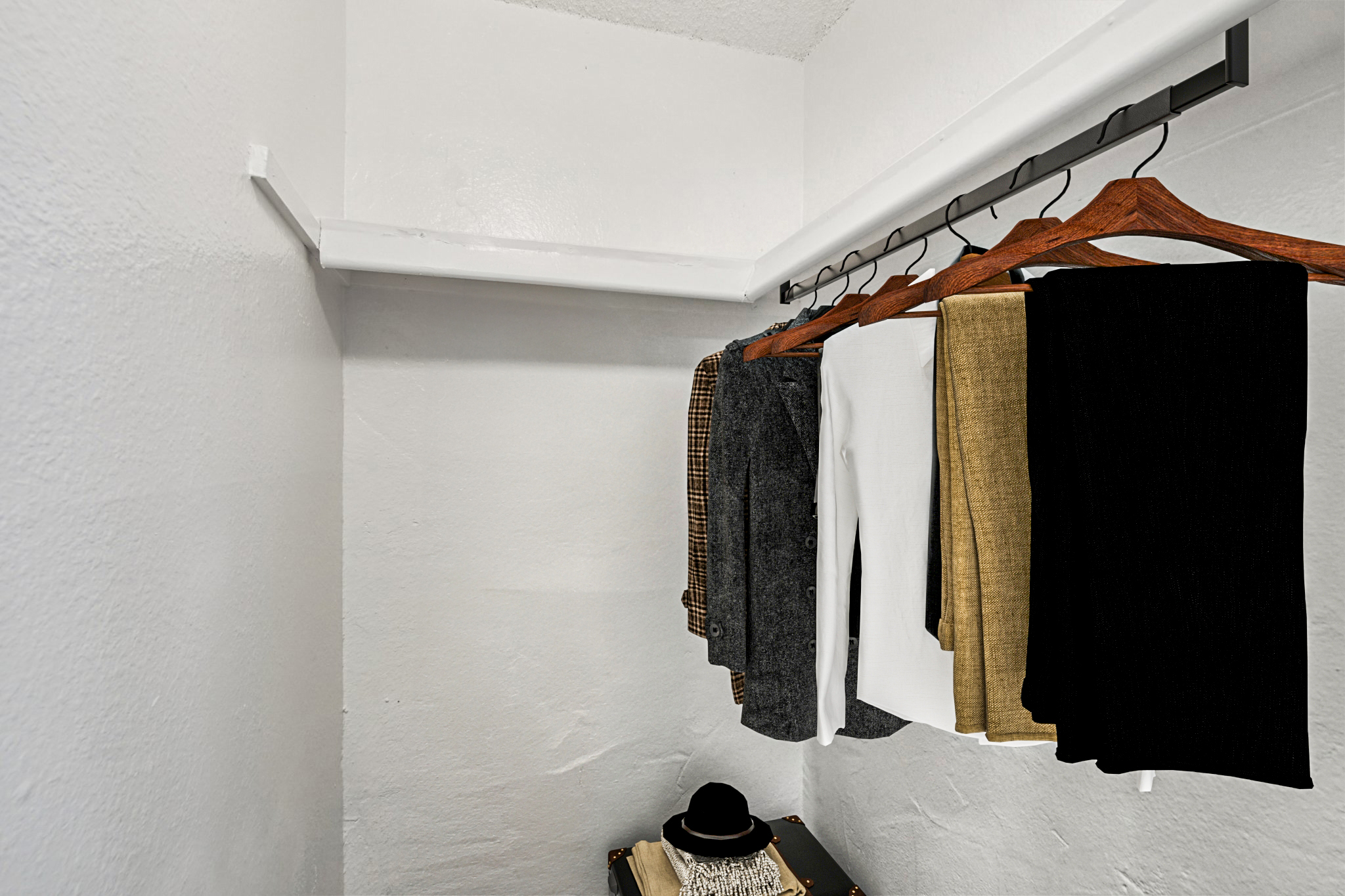 A minimalist closet scene featuring a row of neatly hung clothing on wooden hangers. The garments include various colors and styles, with a focus on neutral tones such as black, white, beige, and gray. A small black hat rests on a decorative surface beneath the clothing, against a plain white wall.
