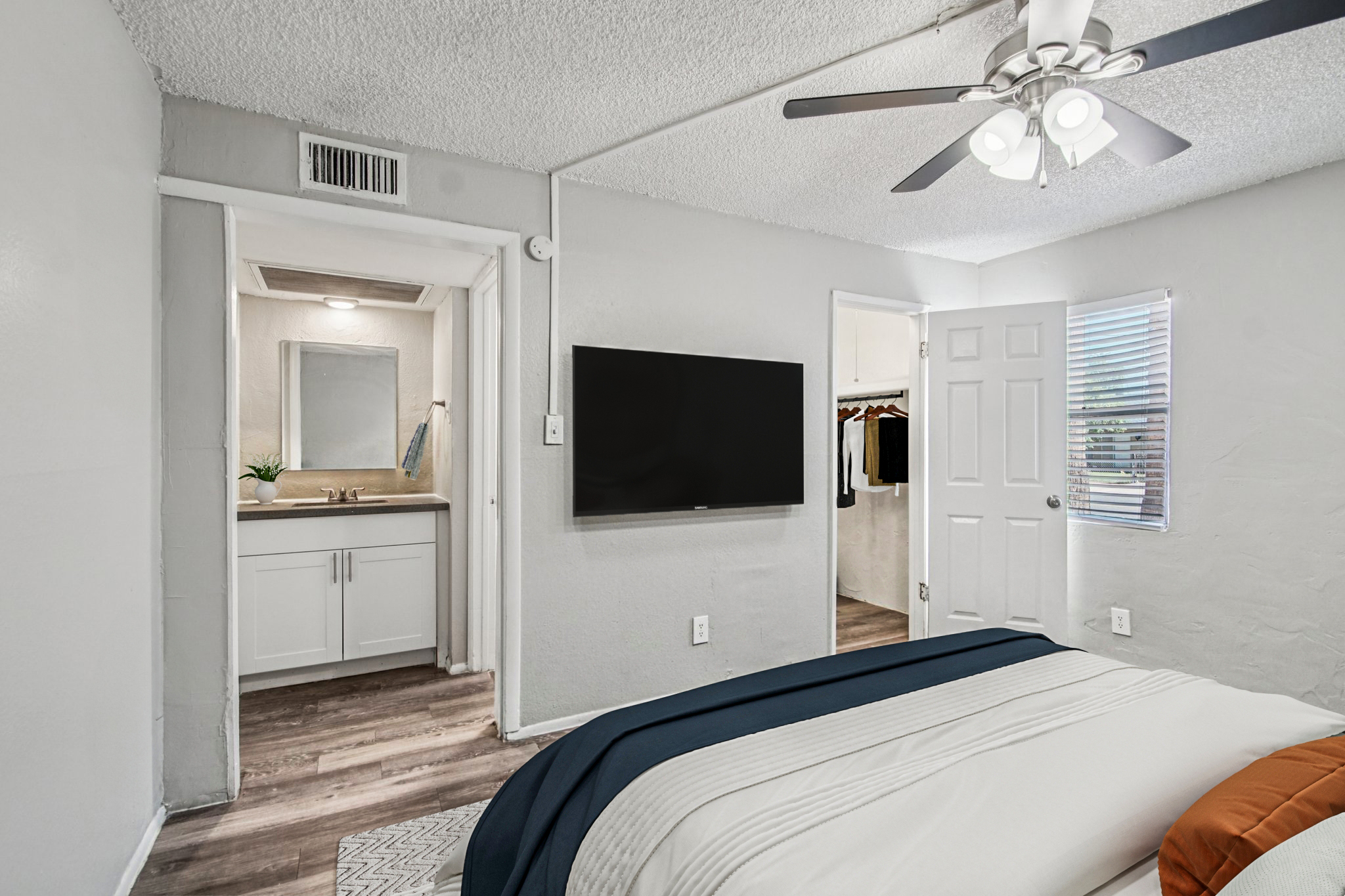 A cozy bedroom featuring a bed with white linens, a ceiling fan above, and a wall-mounted TV. A doorway leads to a bathroom area with a mirror and sink. Natural light enters through a window, and there's a closet visible in the adjacent room. The walls are painted light gray, creating a calm atmosphere.