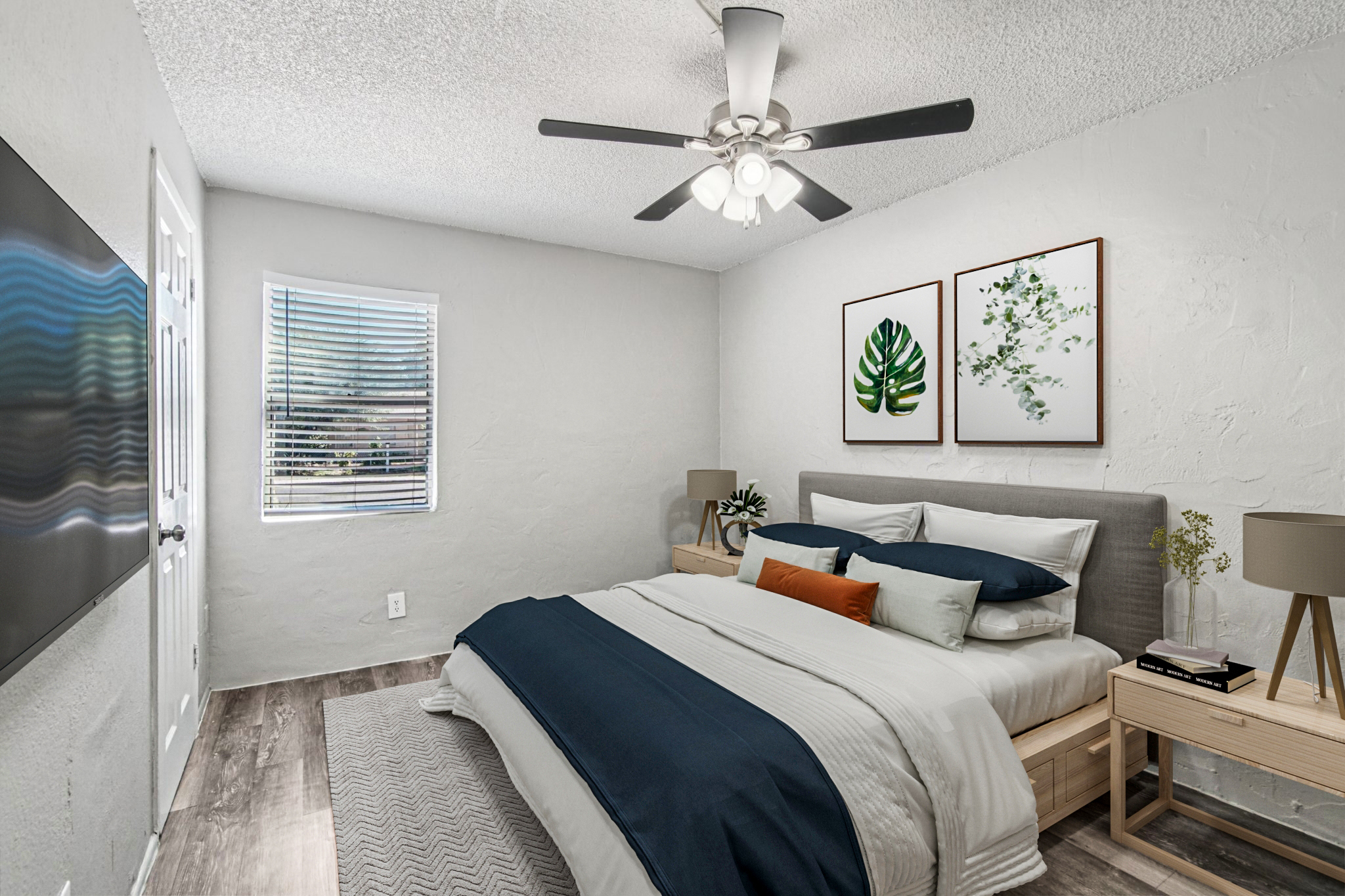 A cozy bedroom featuring a bed with white and navy bedding, accented by decorative pillows. There are two framed botanical prints on the wall, a ceiling fan, and a closet door. Natural light comes through a window with blinds, and there is a small nightstand with a lamp on either side of the bed.