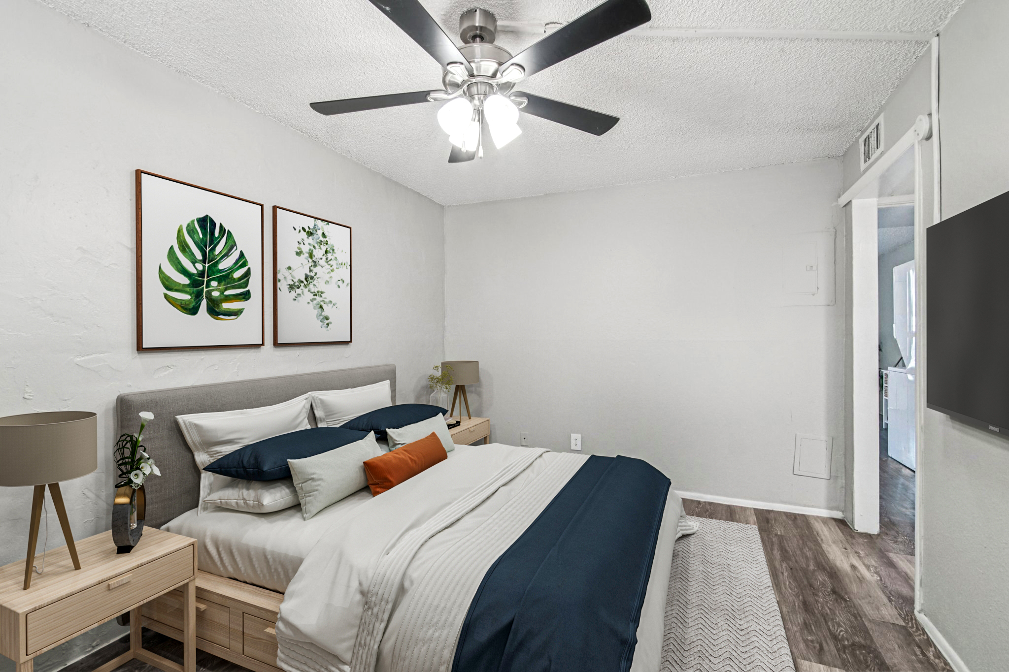 A cozy bedroom featuring a large bed with navy and white bedding, decorative pillows, and two nightstands with lamps. The walls are painted light gray, adorned with two framed botanical prints. A ceiling fan provides airflow, and a doorway leads to another room, while a wall-mounted TV is visible.