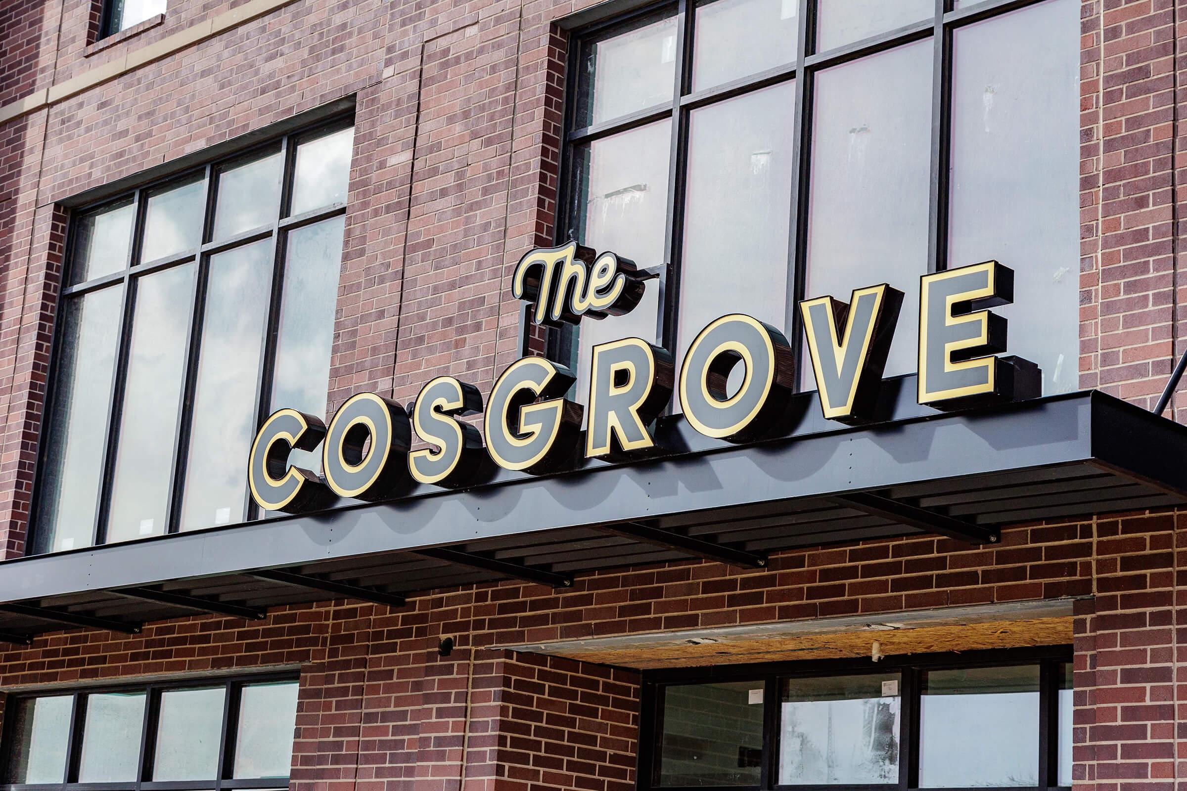 Sign reading "The COSGROVE" in bold, decorative lettering on a building facade made of dark brick, with large windows above. The sign is prominent and stylish, suggesting a welcoming entrance to a business or establishment.