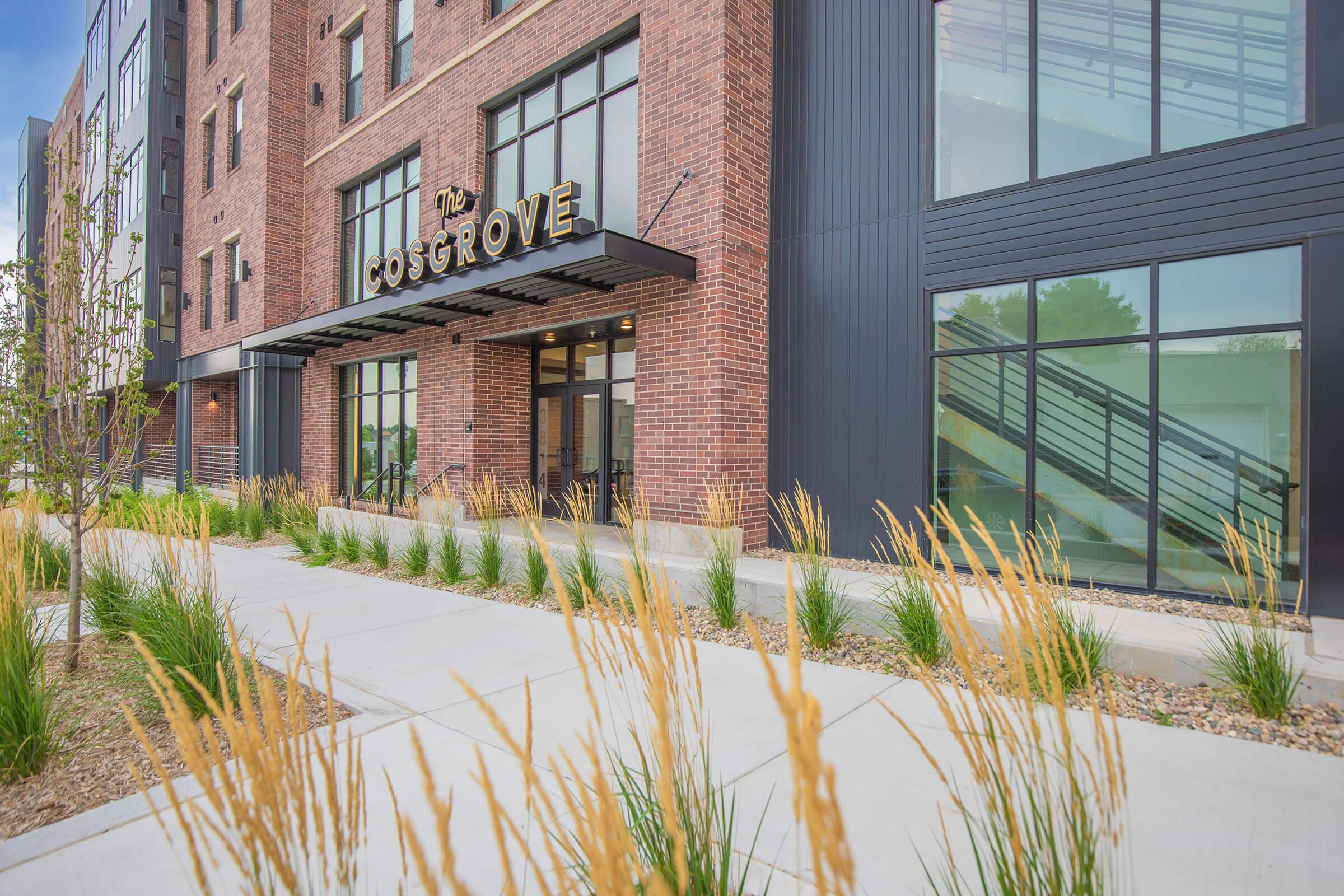 Exterior view of a modern building named "The Cosgrove," featuring red brick facades, large windows, and a welcoming entrance. The landscaping includes ornamental grasses along a paved pathway, leading to the building's entrance.