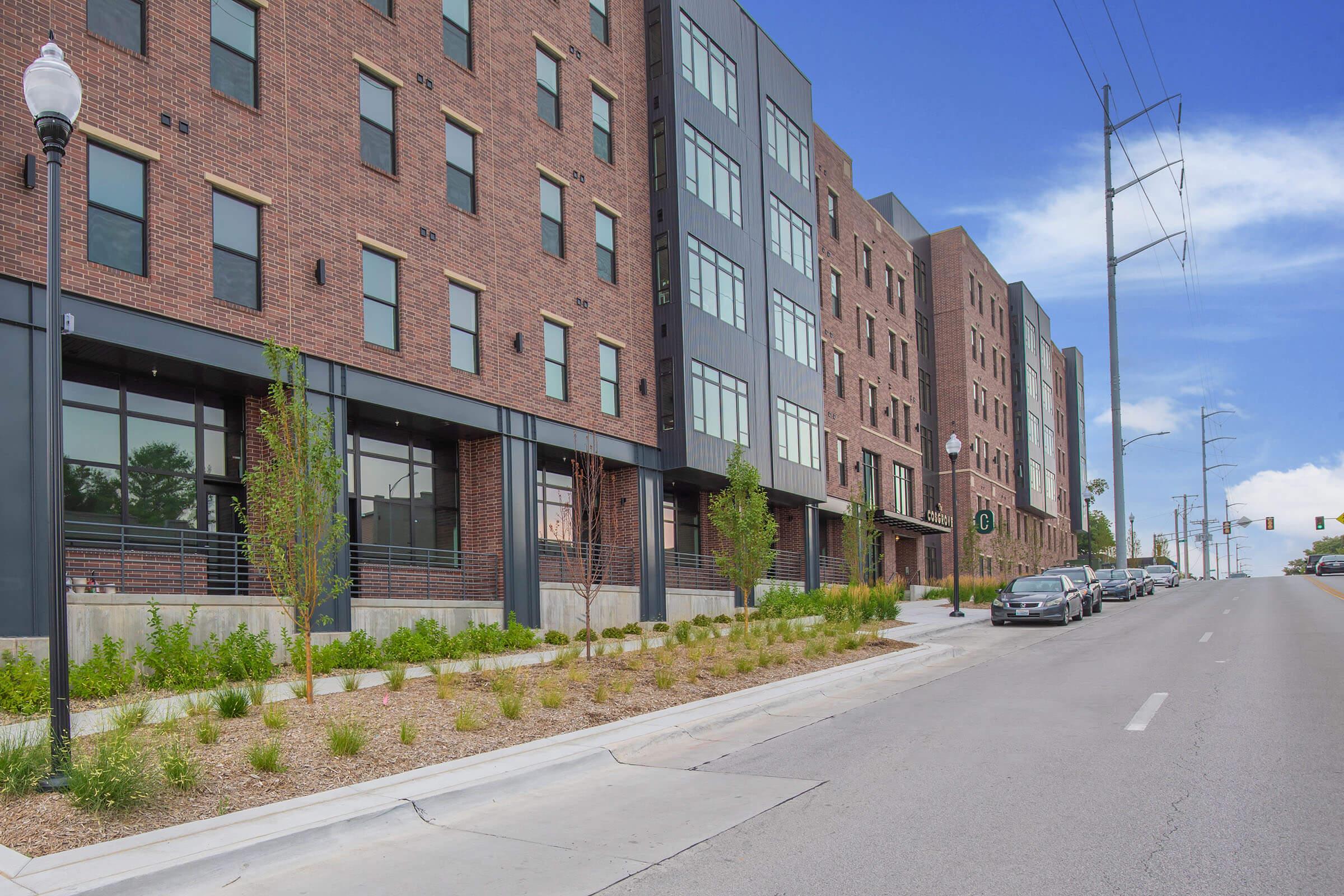 A modern brick building with multiple stories, featuring large windows and a landscaped front area. The street is lined with small trees and shrubs, and parked cars are visible along the side. Bright blue sky in the background enhances the urban setting.