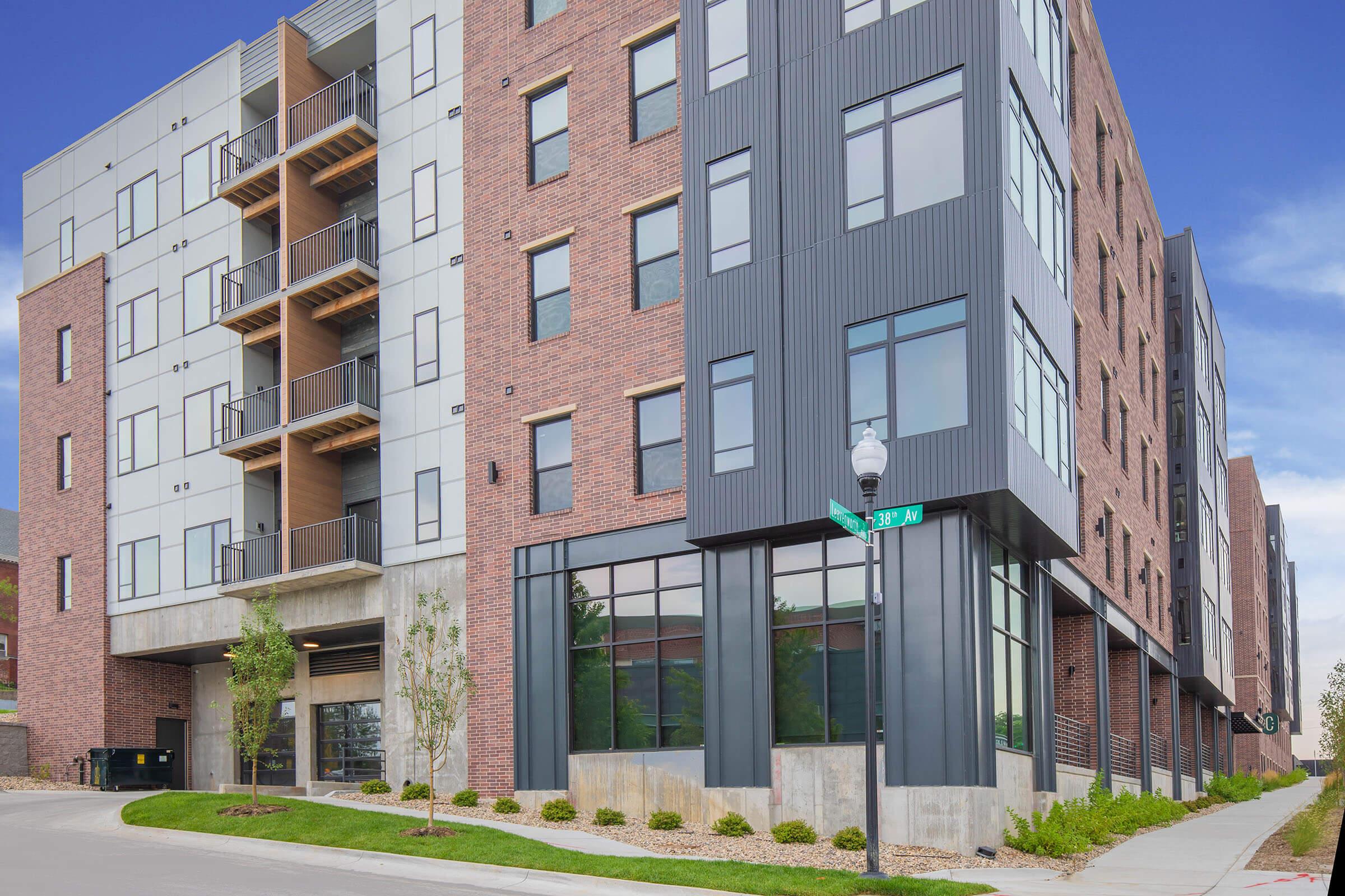 Modern multi-story residential building with a mix of brick and metal facade. Features large windows and balconies on the upper levels, and a concrete ground floor. Situated on a corner lot with landscaping, streetlights, and a clear blue sky in the background.