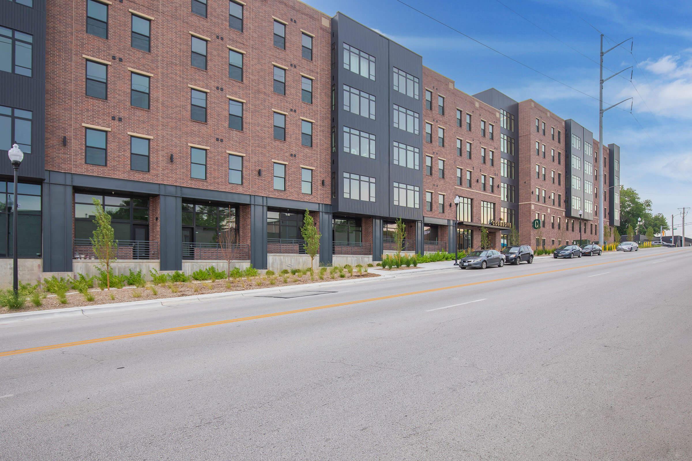 A modern brick apartment building with large windows, featuring several floors and landscaping along the sidewalk. A road runs in front with parked cars and streetlights. The scene is bright and showcases urban living.
