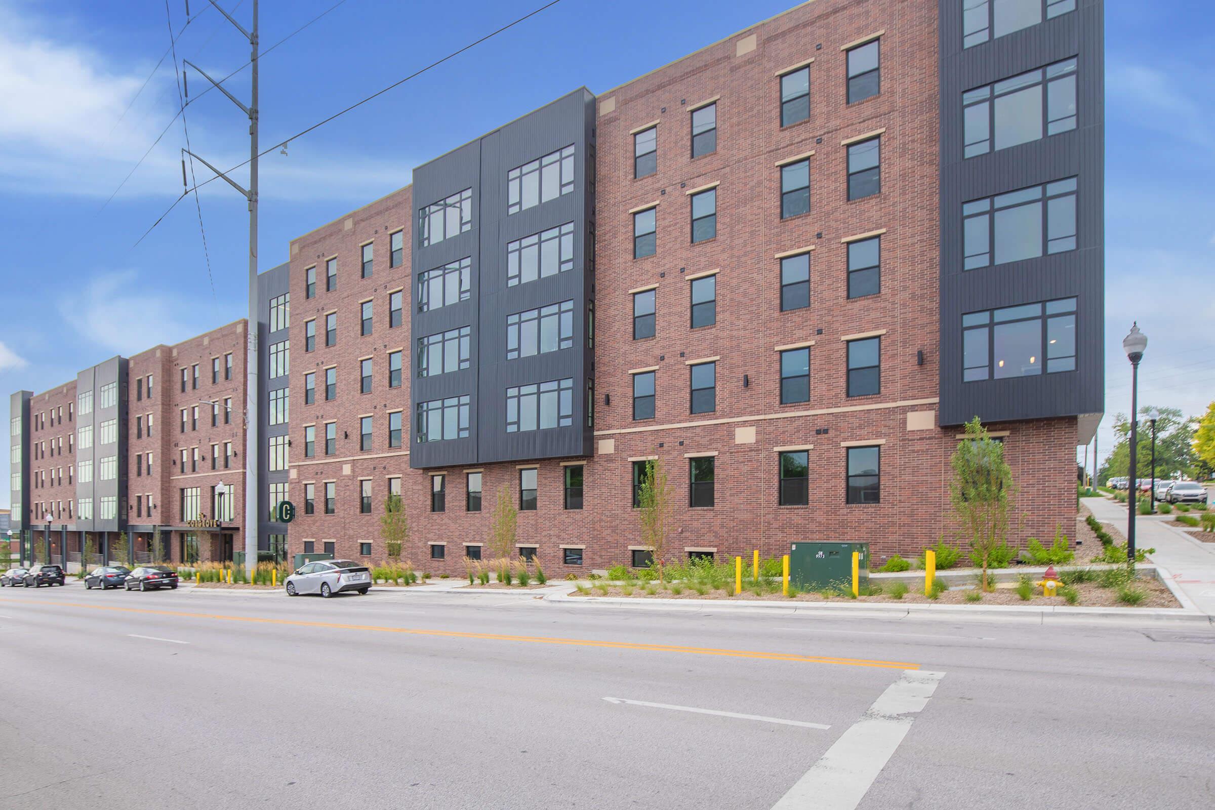 A modern brick building with multiple floors featuring large windows, located along a street. There are cars parked on the road, and greenery surrounding the base of the building. The design combines traditional brickwork with contemporary materials, and it is situated in an urban environment.