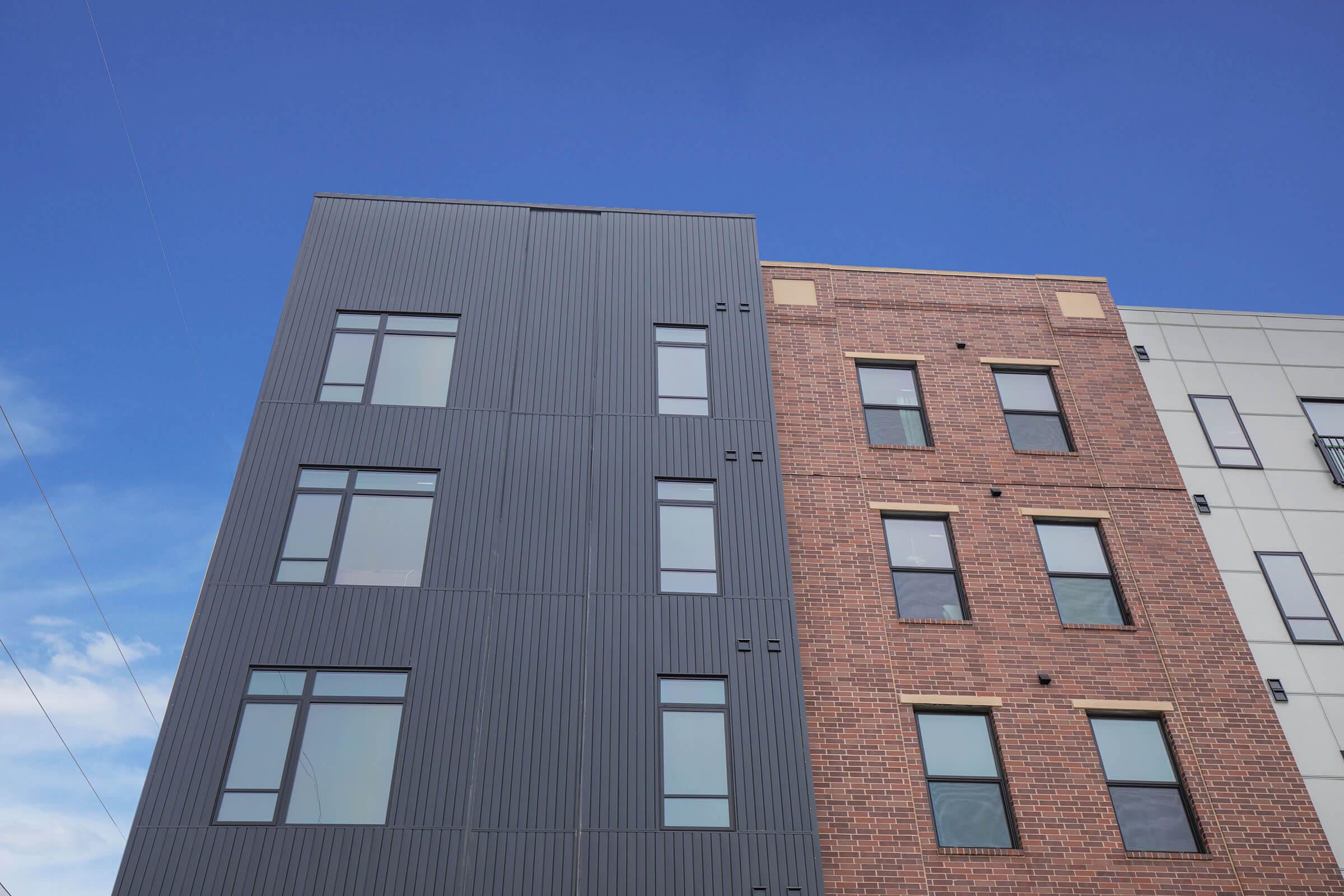 Modern building facade featuring two distinct styles: one side has a sleek, dark, corrugated exterior, while the other side showcases a traditional red brick design with large windows. The background includes a clear blue sky.