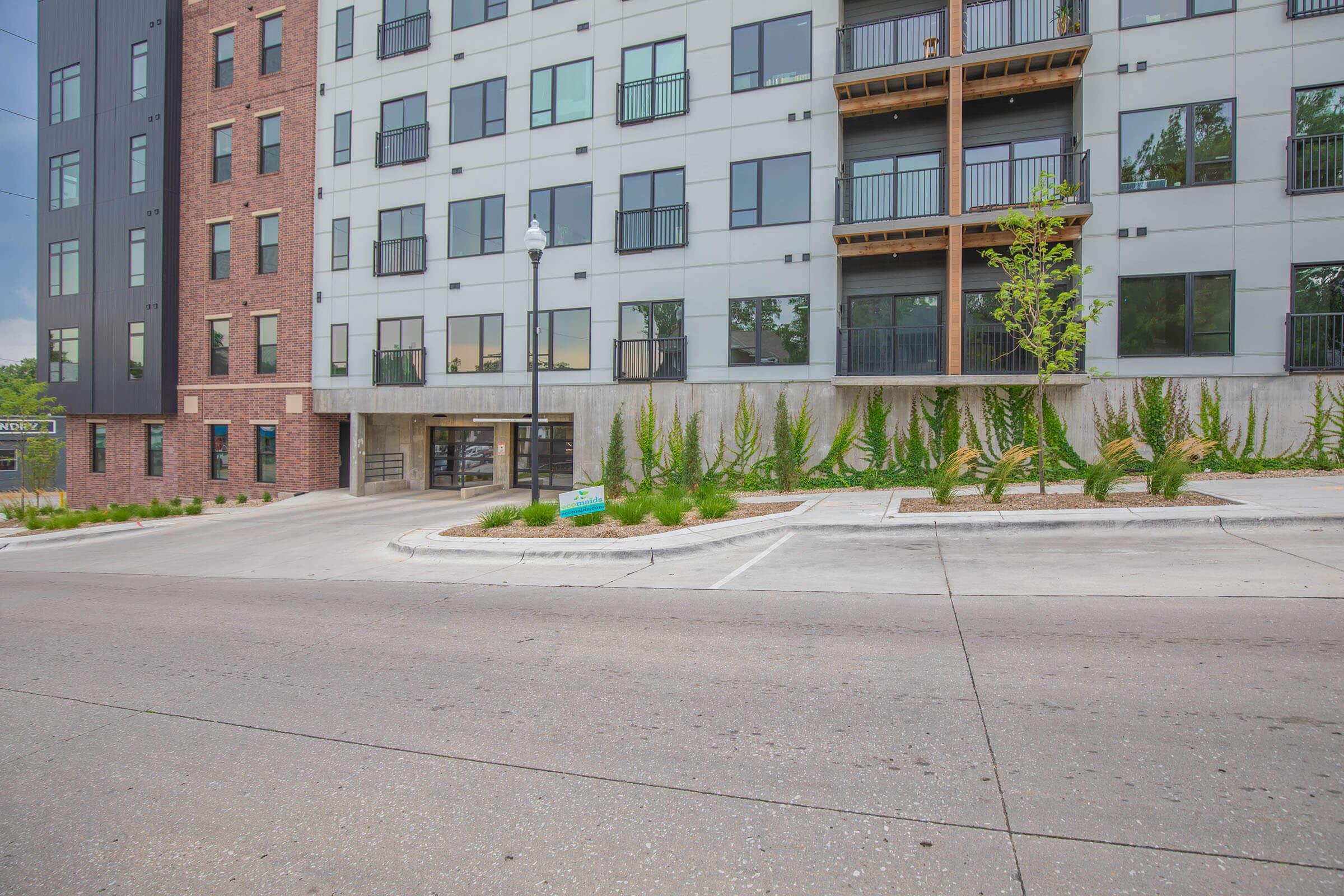 Modern apartment building featuring a mix of brick and gray exterior with large windows. The building has multiple floors, balconies, and landscaping along the entrance. A parking area is visible in front, with a smooth concrete surface and decorative plants lining the driveway.