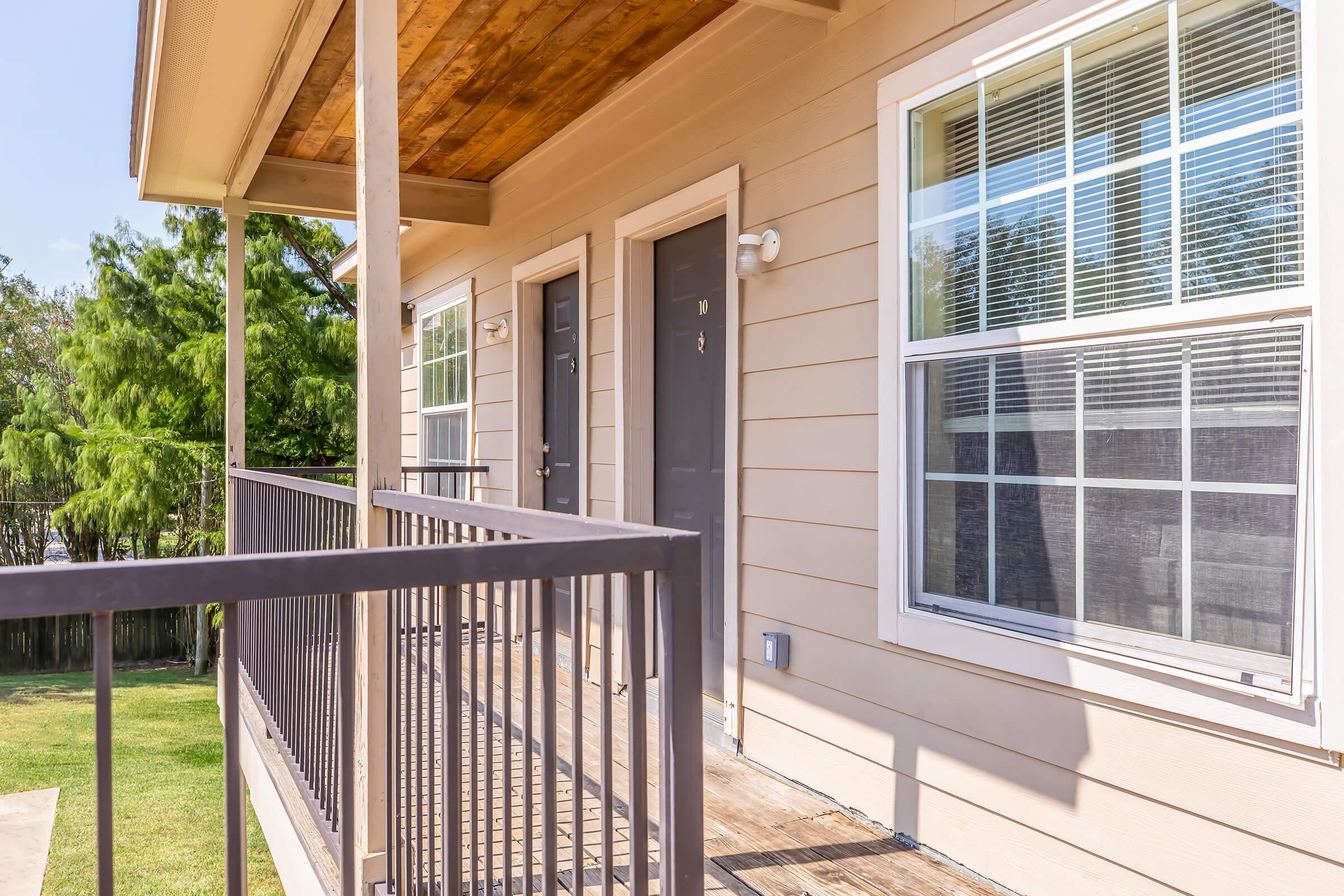 Exterior view of a building with a porch and two doorways. The porch features a wooden railing, and there are large windows beside the doors. The setting is sunny, with greenery visible in the background, indicating a pleasant outdoor environment.