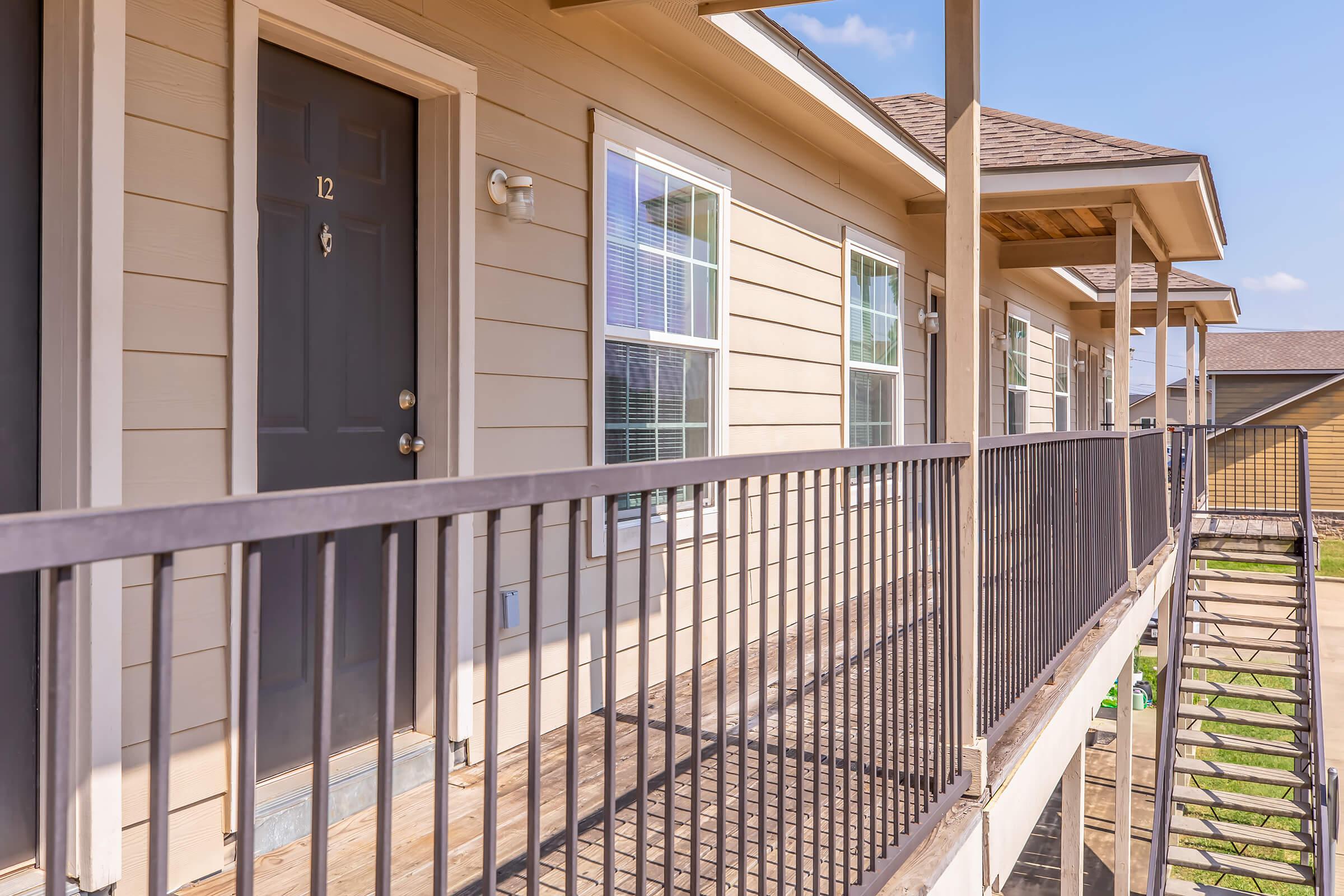 Exterior view of an apartment building showcasing a second-floor balcony with a black metal railing. The image features a brown door marked "12" and several windows with white frames, providing a bright and welcoming atmosphere against a clear blue sky.