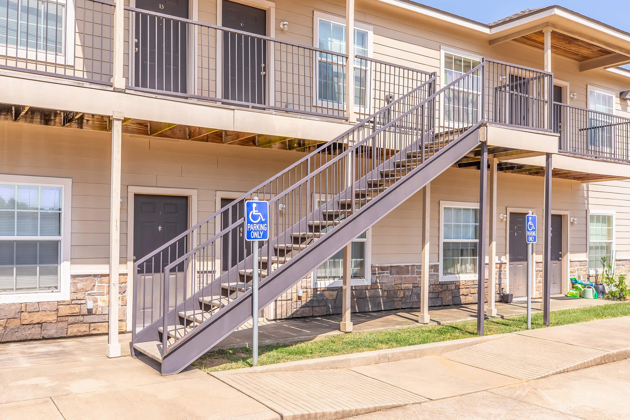 Exterior view of a two-story apartment building featuring a set of metal stairs leading to the second floor. There are two designated "Handicap Parking Only" signs visible. The building has a stone lower exterior with beige siding and multiple windows. A green lawn is in front of the stairs.