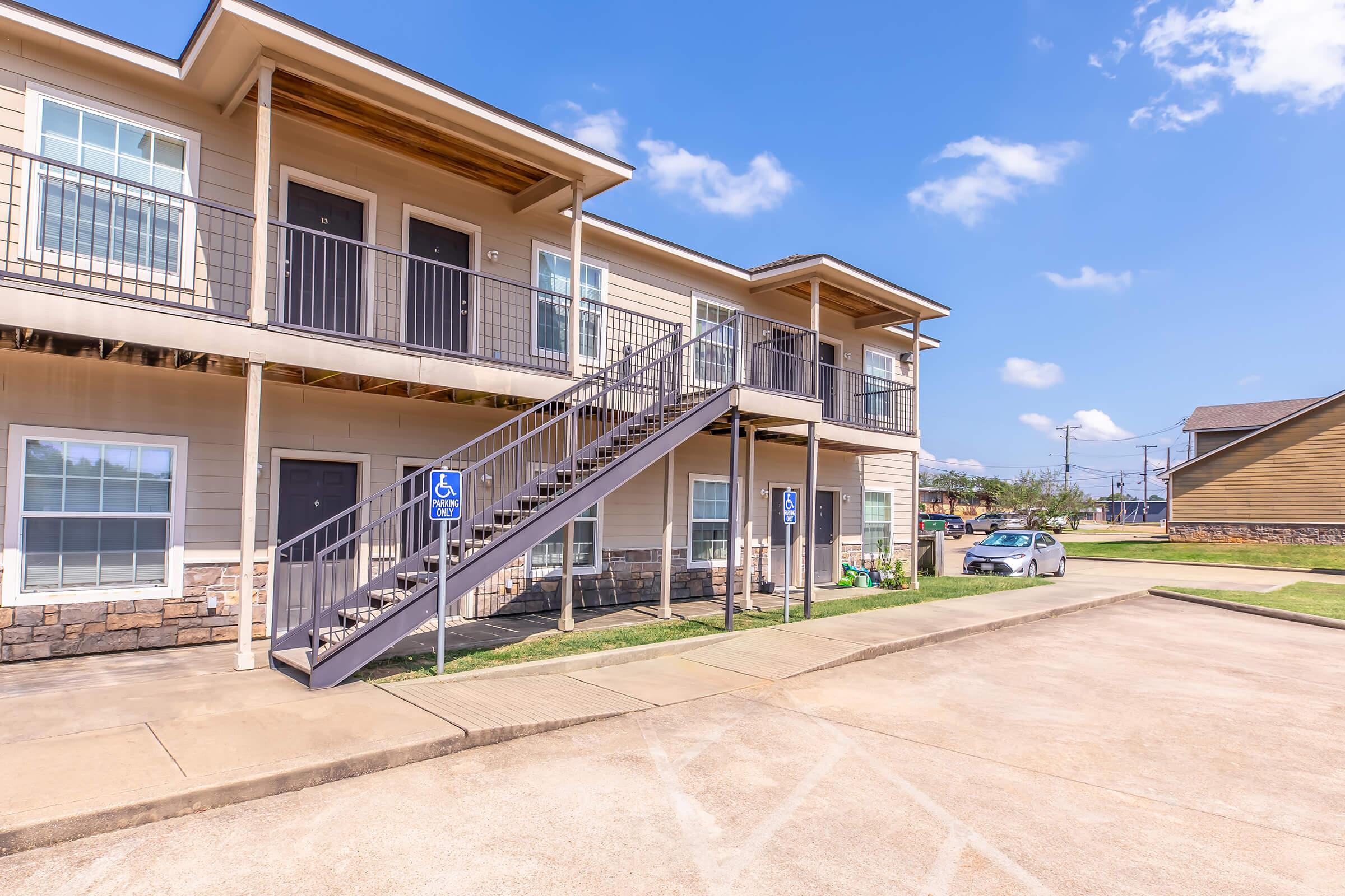 A two-story apartment building featuring a staircase leading to the upper level. The building has multiple doors, parking spaces in front, and is surrounded by green grass. Blue sky with a few clouds is visible above, and there are accessible parking spots in front of the building.