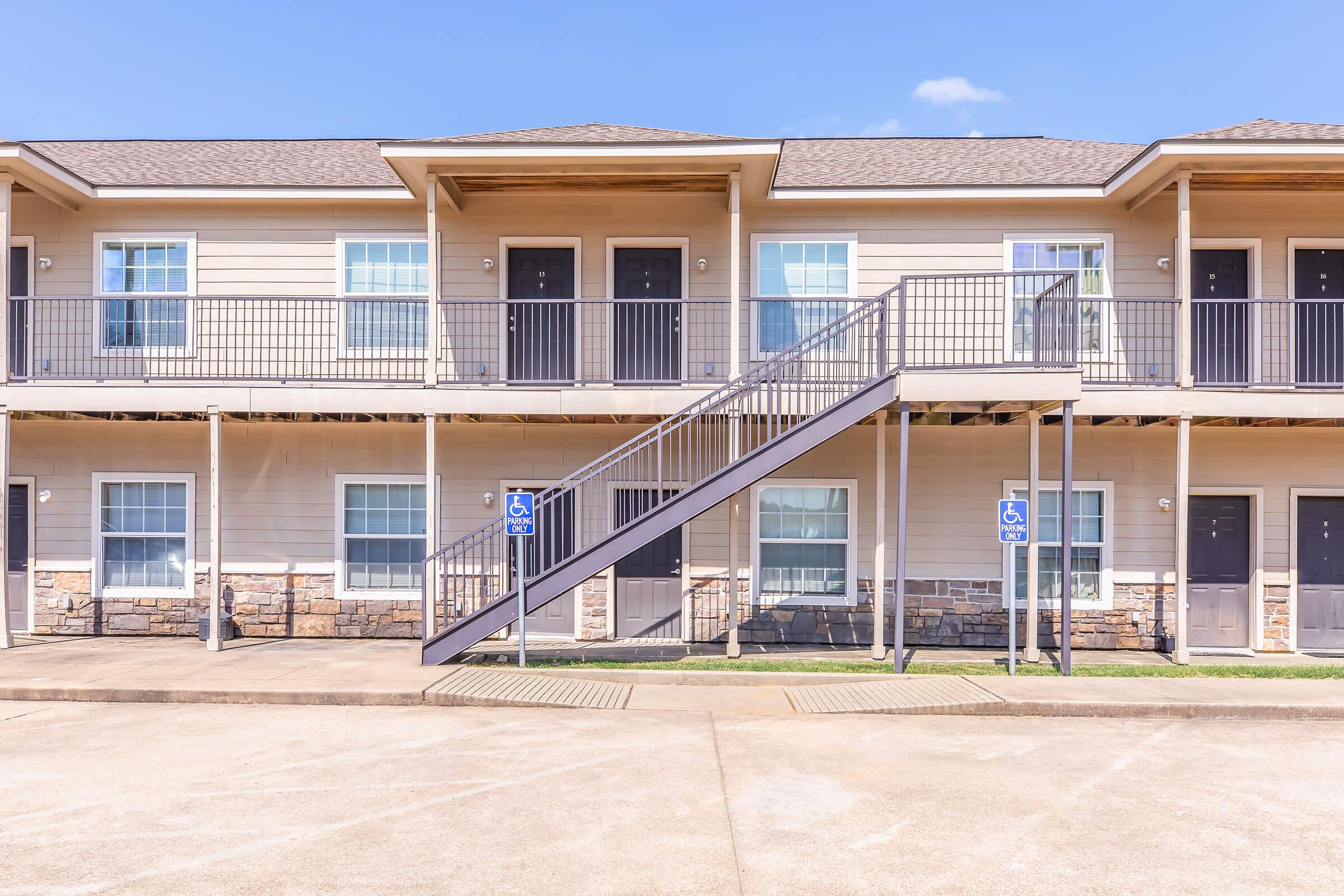 Two-story apartment building with a light-colored exterior and stone accents. The building features a staircase leading to the upper level, with several entrances visible. The ground level includes designated parking spaces, and the sky is clear and blue, enhancing the overall bright and inviting appearance.