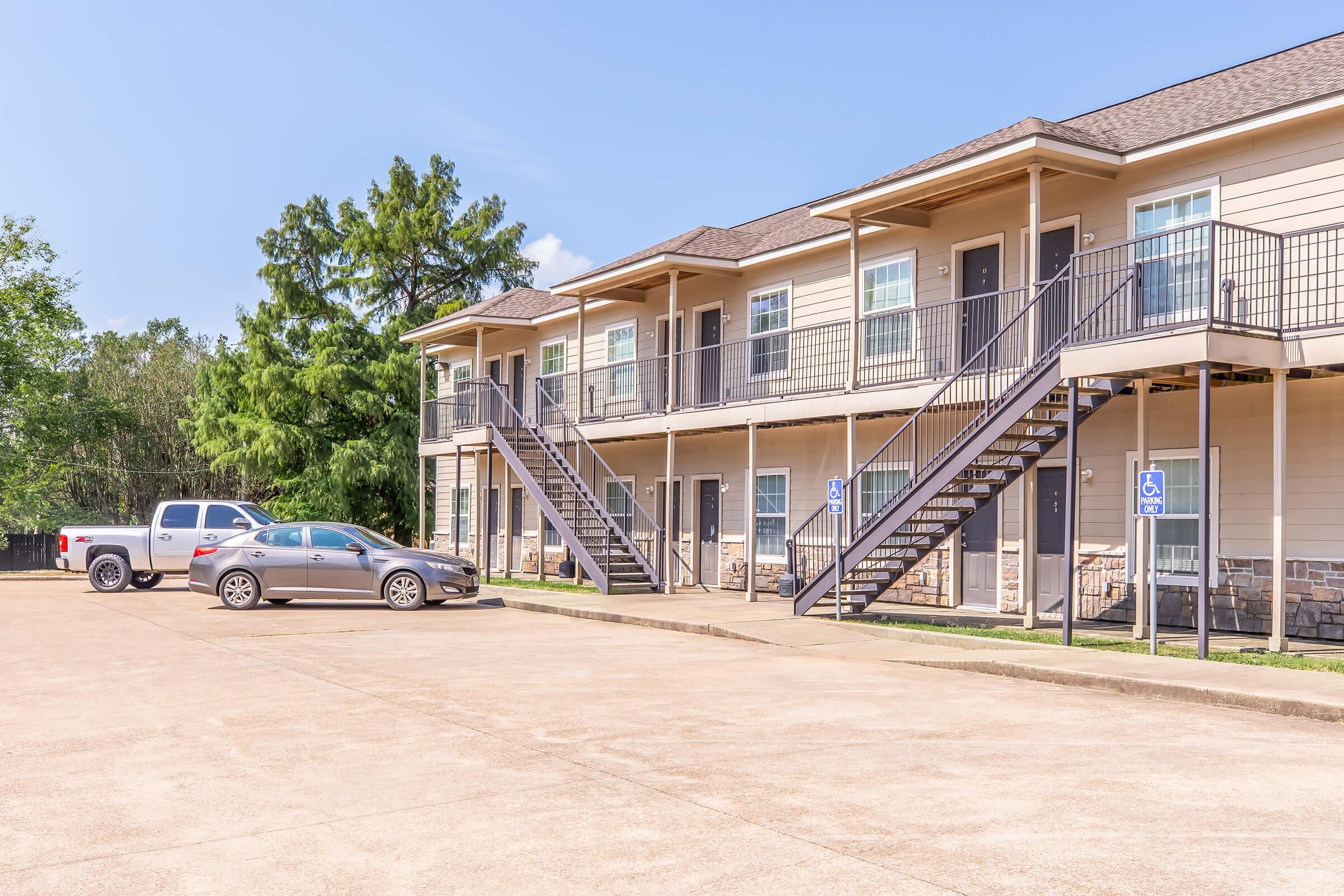 A view of a two-story apartment building with multiple units, featuring balconies and metal staircases. A paved parking lot in front contains two parked cars. Surrounding the building are trees and a clear blue sky.