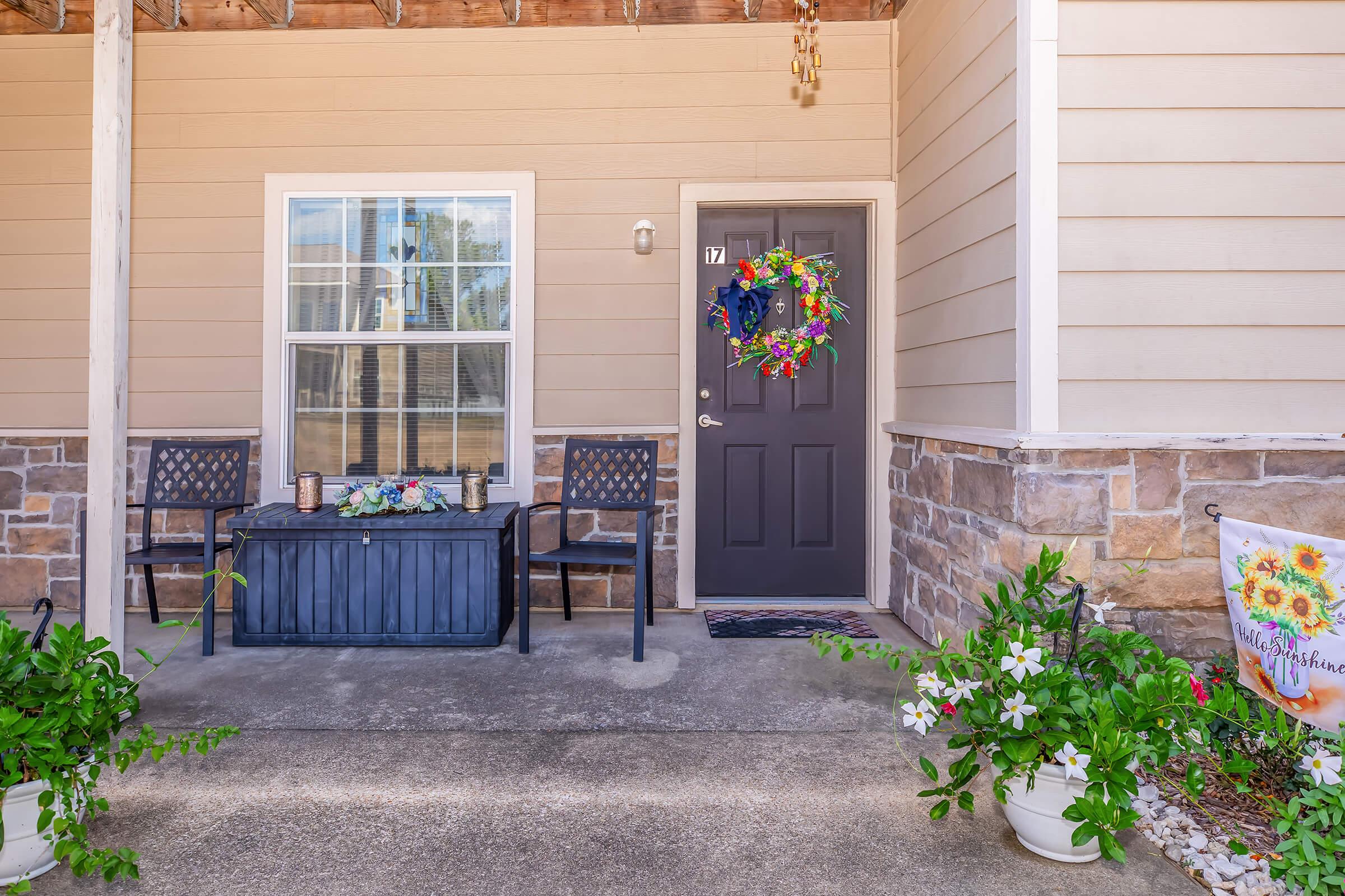 A cozy porch featuring a dark front door with the number 17, a decorative wreath, and two chairs beside a dark coffee table. Potted plants with white flowers are placed at the base of the porch, and a banner with sunflowers hangs to the right, adding a welcoming touch to the entrance.