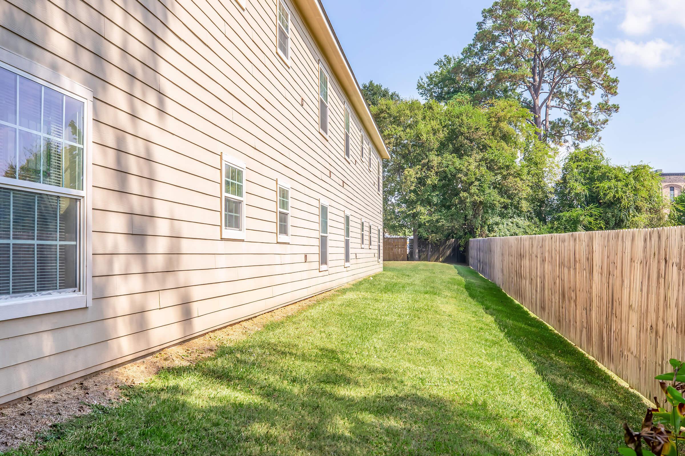 A narrow side yard of a light-colored house featuring a well-maintained lawn, with trees and a wooden fence visible in the background under a clear blue sky. The house has several windows on its side, and the area is well-lit by natural sunlight.