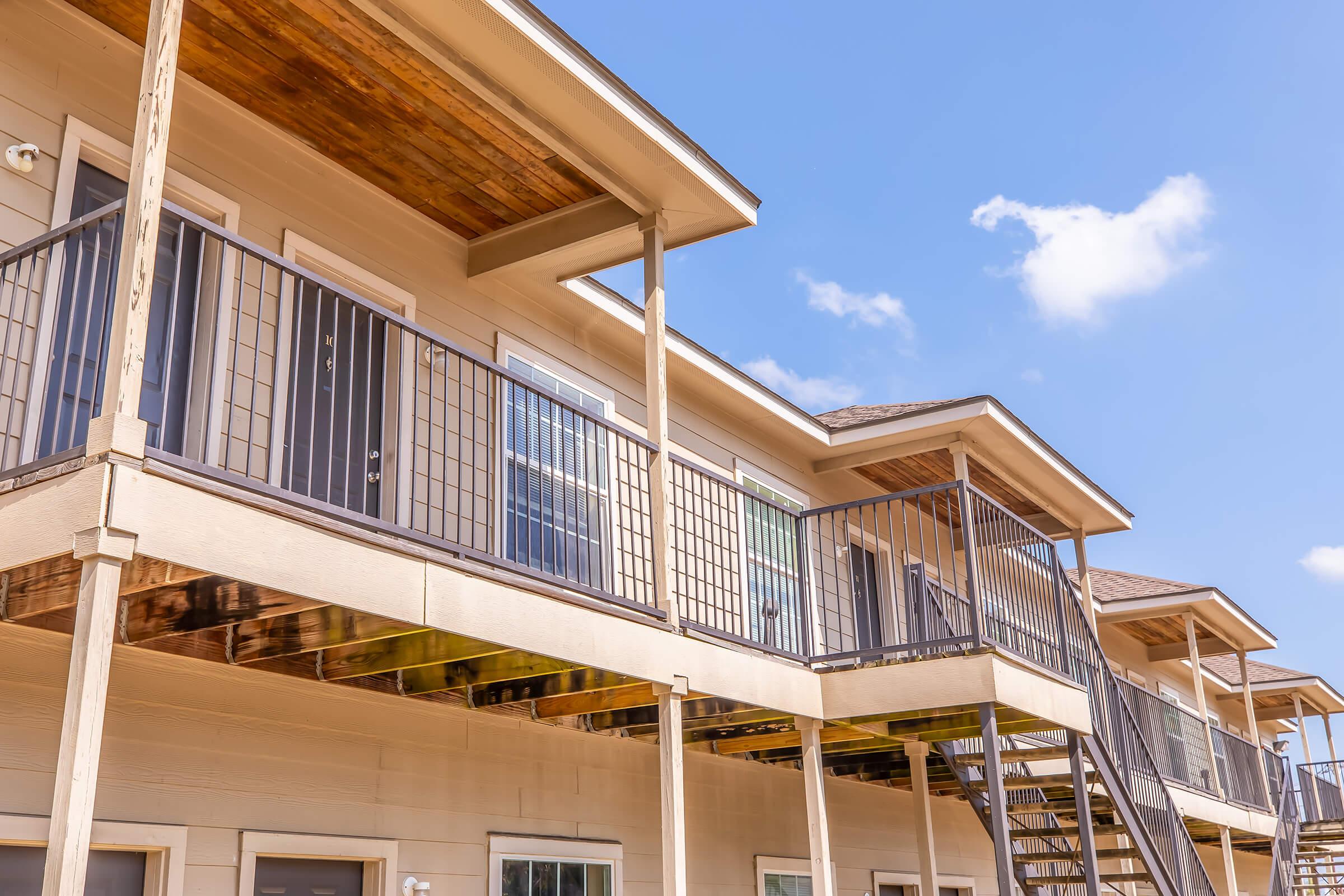 A view of two stories of an apartment building, featuring multiple balconies with railings. The exterior is painted in neutral colors, and there are several window panes visible. The sky above is blue with a few clouds, creating a bright, inviting atmosphere.