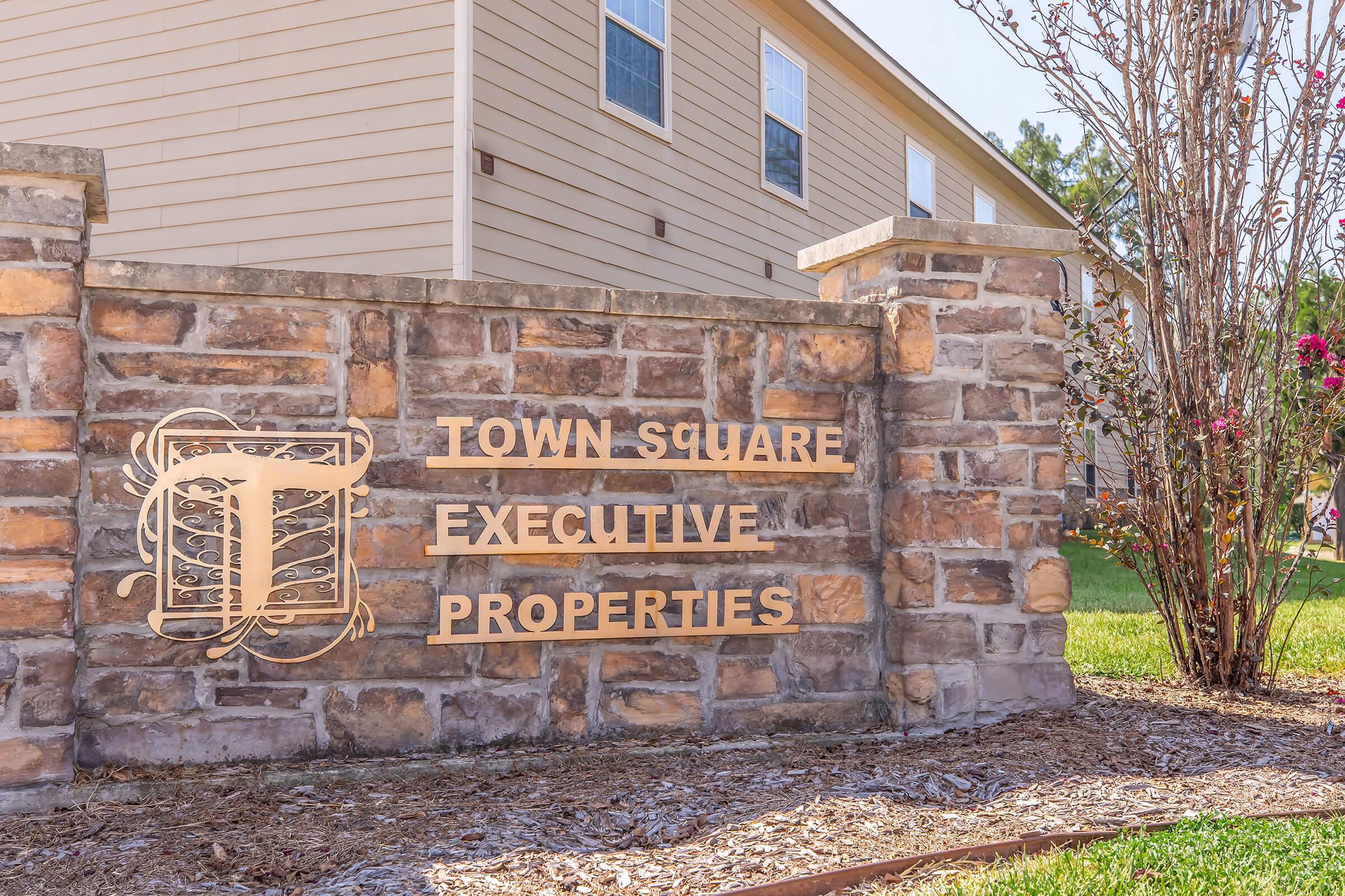 Sign displaying "Town Square Executive Properties" made of gold-colored metal, mounted on a stone wall, surrounded by green grass and a flowering shrub. In the background, a beige building is partially visible.