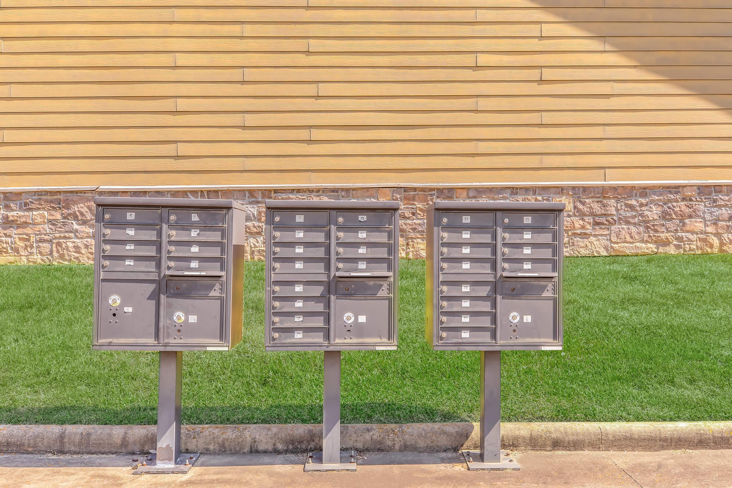 A row of three metal mailboxes mounted on gray poles, positioned in front of a low stone wall and a light-colored wooden facade. The area is bordered by green grass, creating a neat and organized appearance.