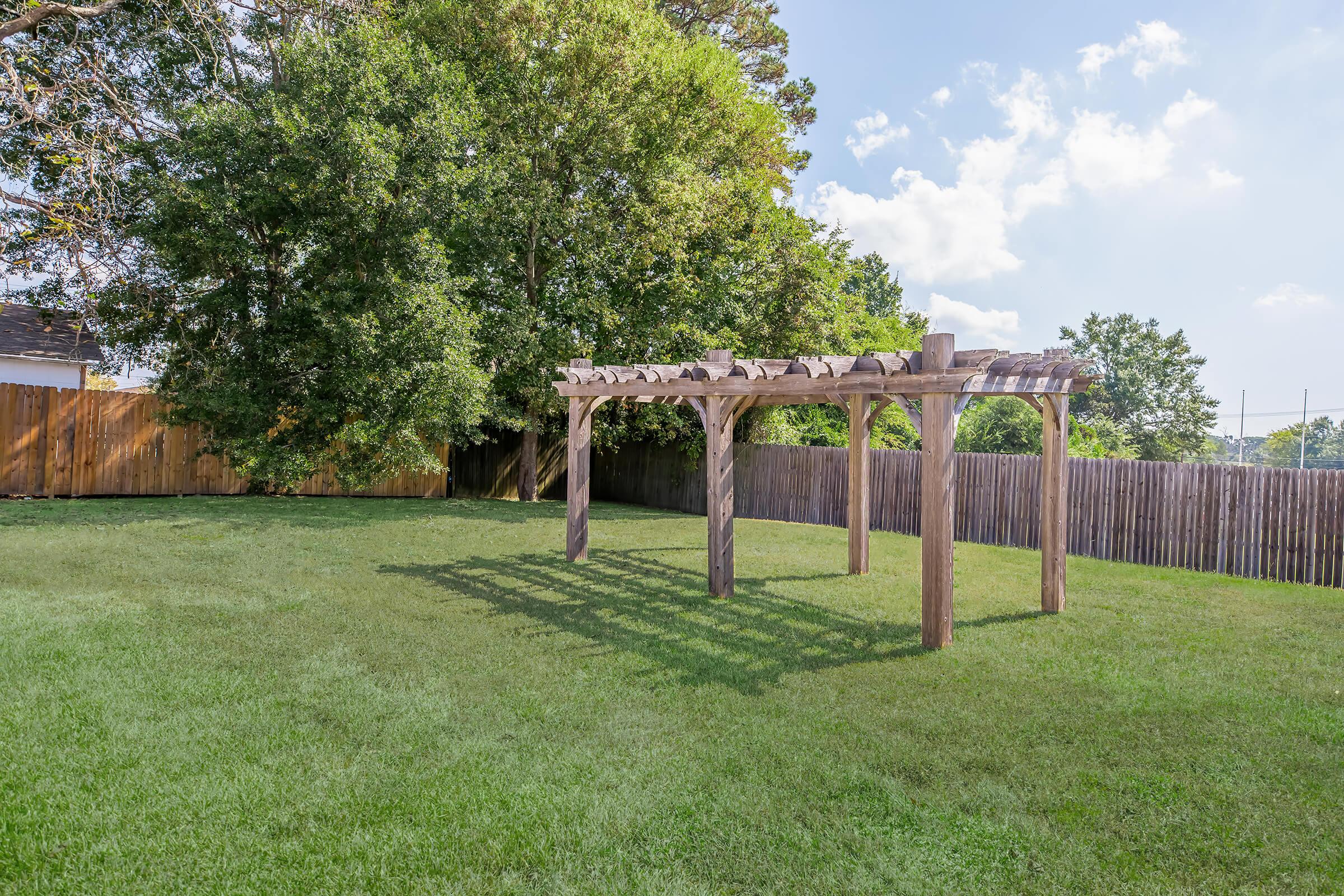 A wooden pergola stands in a grassy backyard surrounded by a wooden fence. Lush trees provide shade in the background, and the sky is partly cloudy, creating a serene outdoor atmosphere.