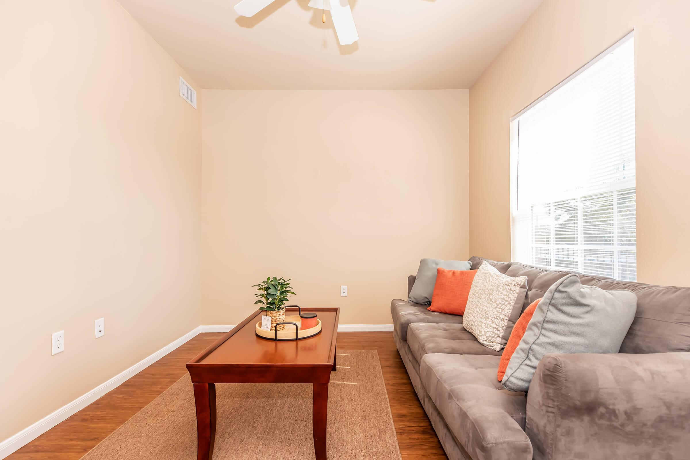 A cozy living room featuring a gray sofa adorned with orange and light blue pillows. In front of the sofa, there's a brown wooden coffee table with a decorative tray and a small plant. The walls are painted beige, and natural light streams in through a window, adding warmth to the space.