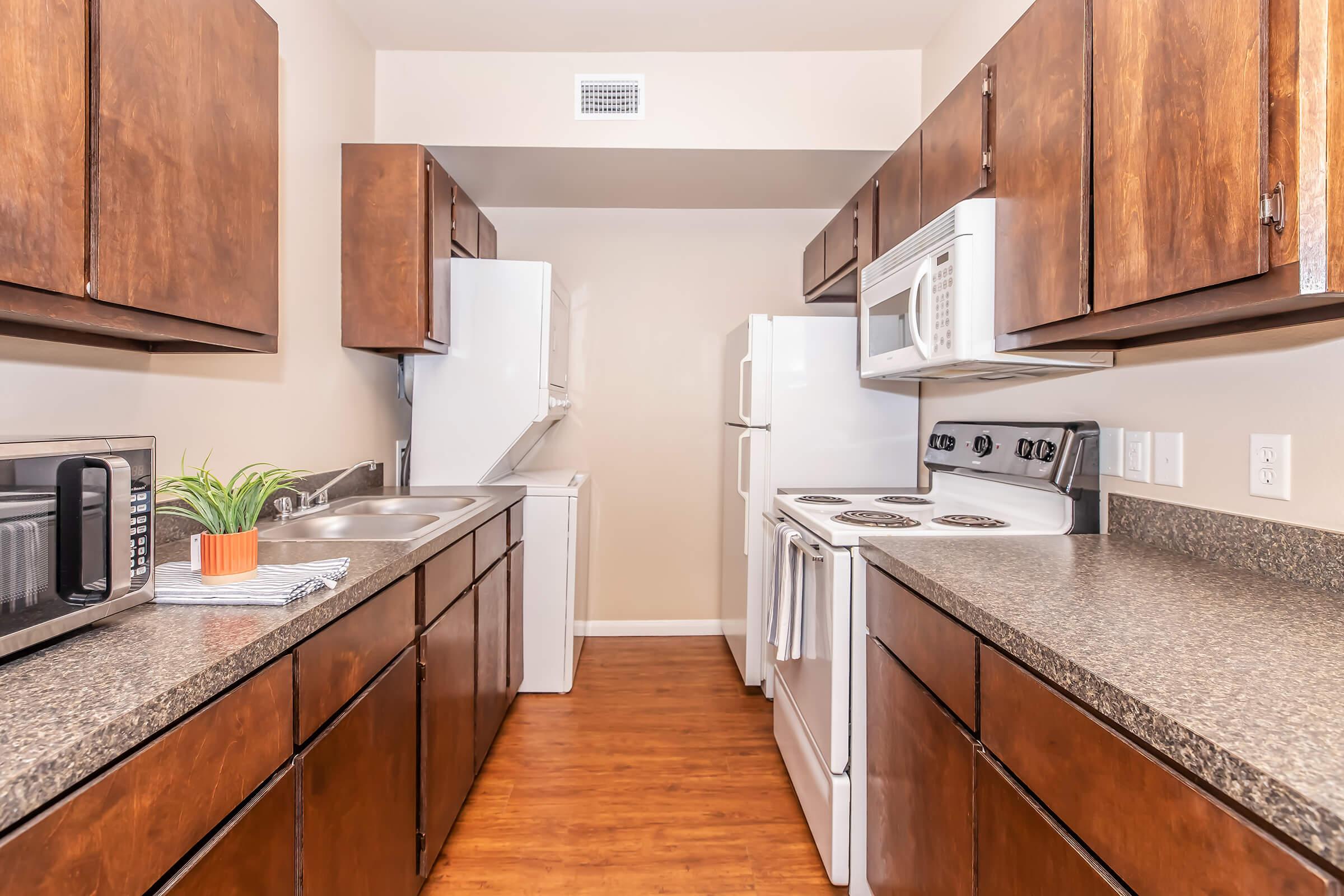 A modern kitchen featuring wood cabinetry, granite countertops, and stainless steel appliances, including a refrigerator, microwave, and stove. The space is well-lit with a neutral wall color and a small potted plant on the counter, creating a clean and organized atmosphere.