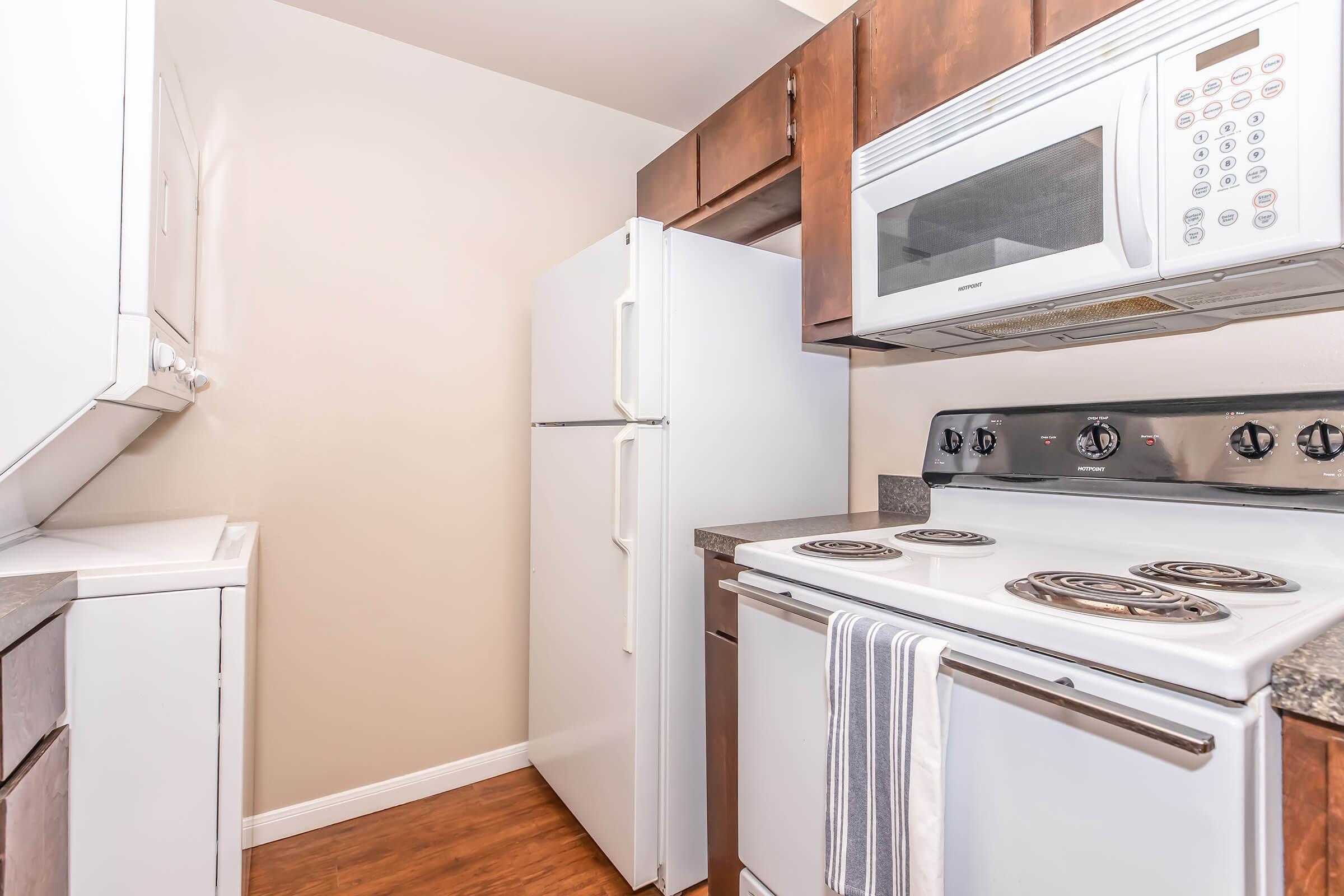 A modern kitchen featuring a white refrigerator, an oven with a stove top, and a microwave above. The cabinets are dark wood, and there is a light-colored wall with a vinyl floor. A kitchen towel is hanging from the oven. The layout is compact and functional.