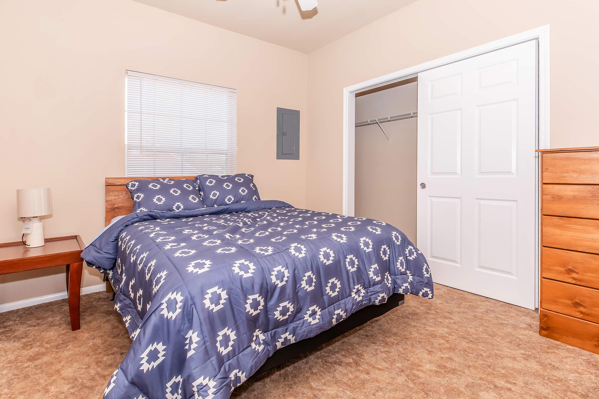 A cozy bedroom featuring a queen-sized bed with blue patterned bedding, a wooden nightstand with a lamp, a dresser, and a closet with sliding doors. Natural light enters through a window, complementing the neutral wall color and carpeted floor.