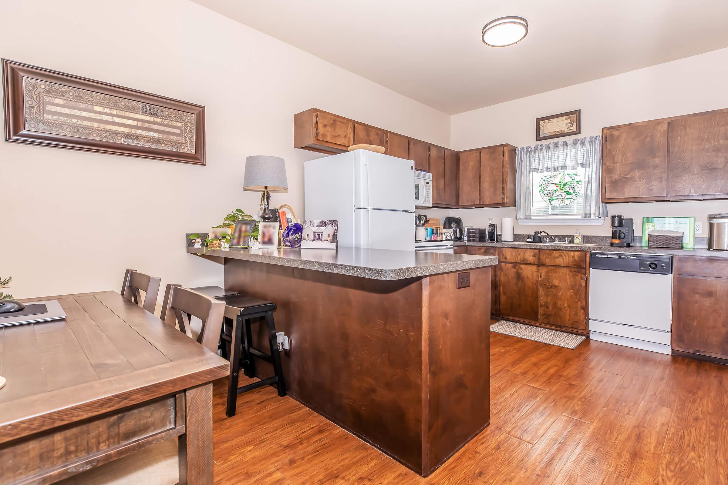 A modern kitchen featuring wooden cabinets, a white refrigerator, and a black dishwasher. There is a small dining table and chairs, a countertop with bar stools, and a window with sheer curtains. The flooring is a warm wood tone, creating a cozy atmosphere.