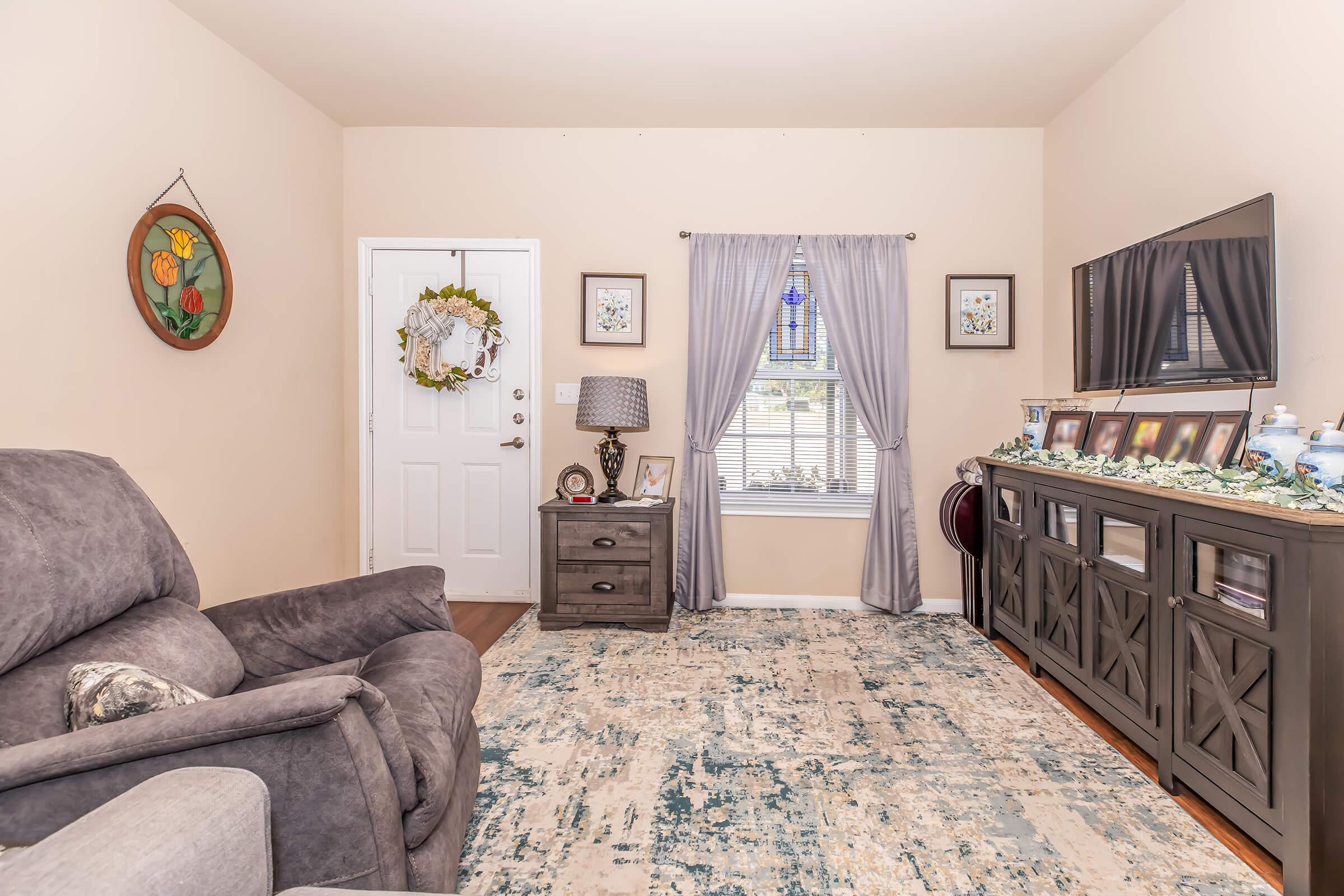 Cozy living room with a gray recliner, a small side table, and a decorated dresser. There's a window with sheer curtains, a door with a wreath, and framed pictures on a media console. Soft-colored carpet adds warmth to the space, creating an inviting atmosphere.
