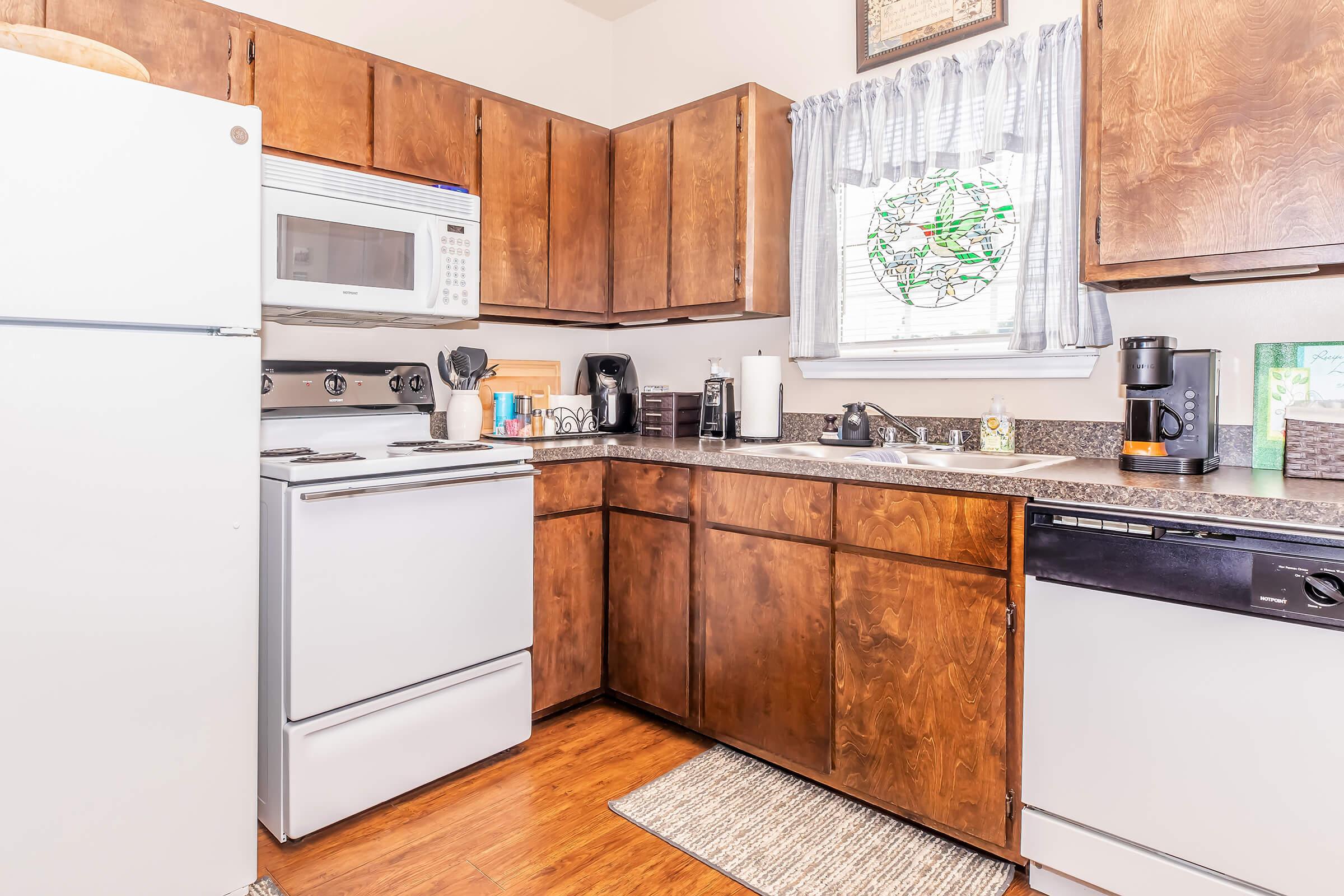 A modern kitchen featuring wooden cabinets, a white refrigerator, a microwave above the stove, and various countertop appliances. The window is dressed with a light curtain, and there's a patterned rug on the floor, creating a warm and inviting atmosphere.
