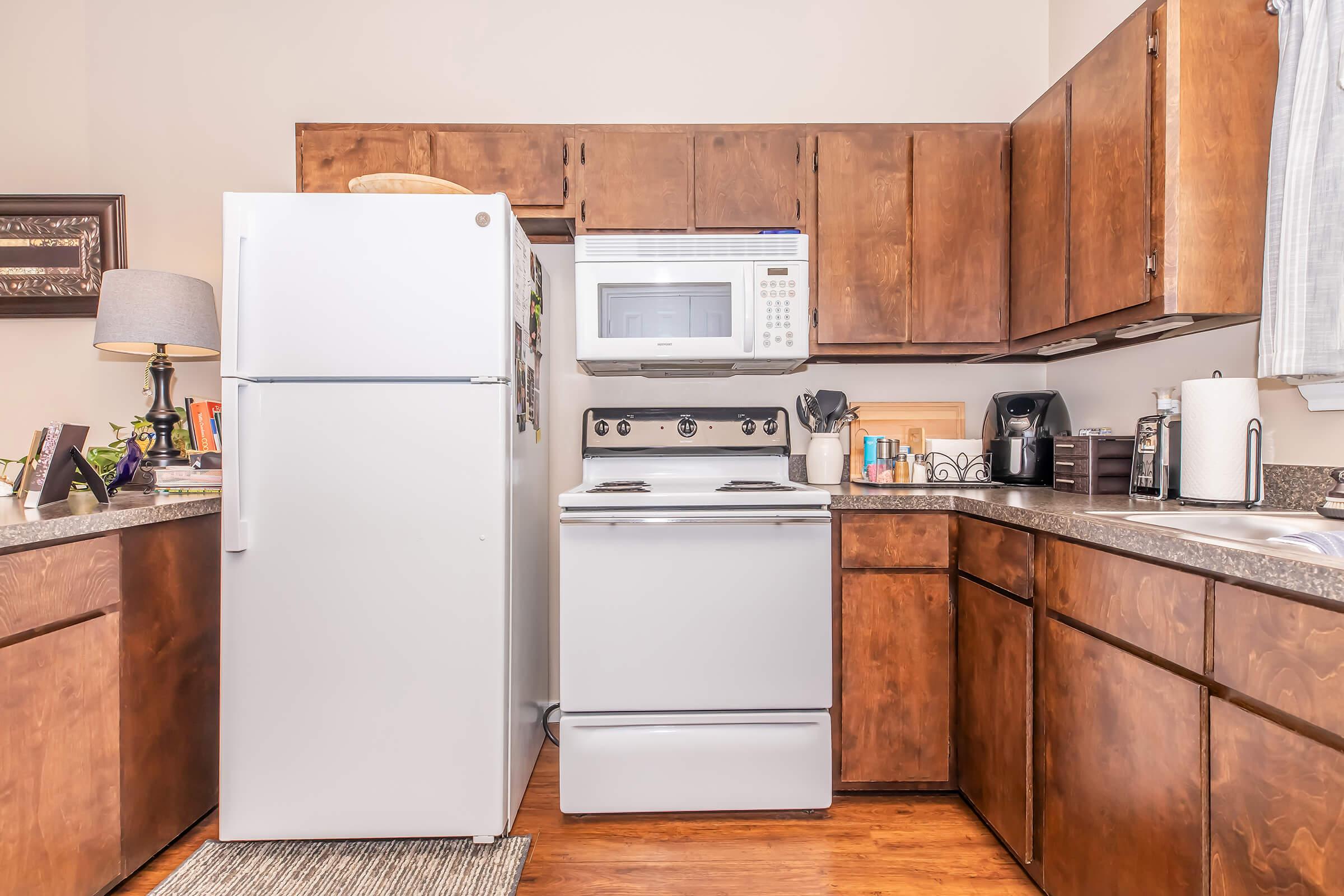 A modern kitchen featuring wooden cabinets, a white refrigerator, a white stove with an oven, and a microwave mounted above. The countertop is adorned with kitchen utensils, a coffee maker, and decorative items. Natural light filters through a window, creating a warm and inviting atmosphere.