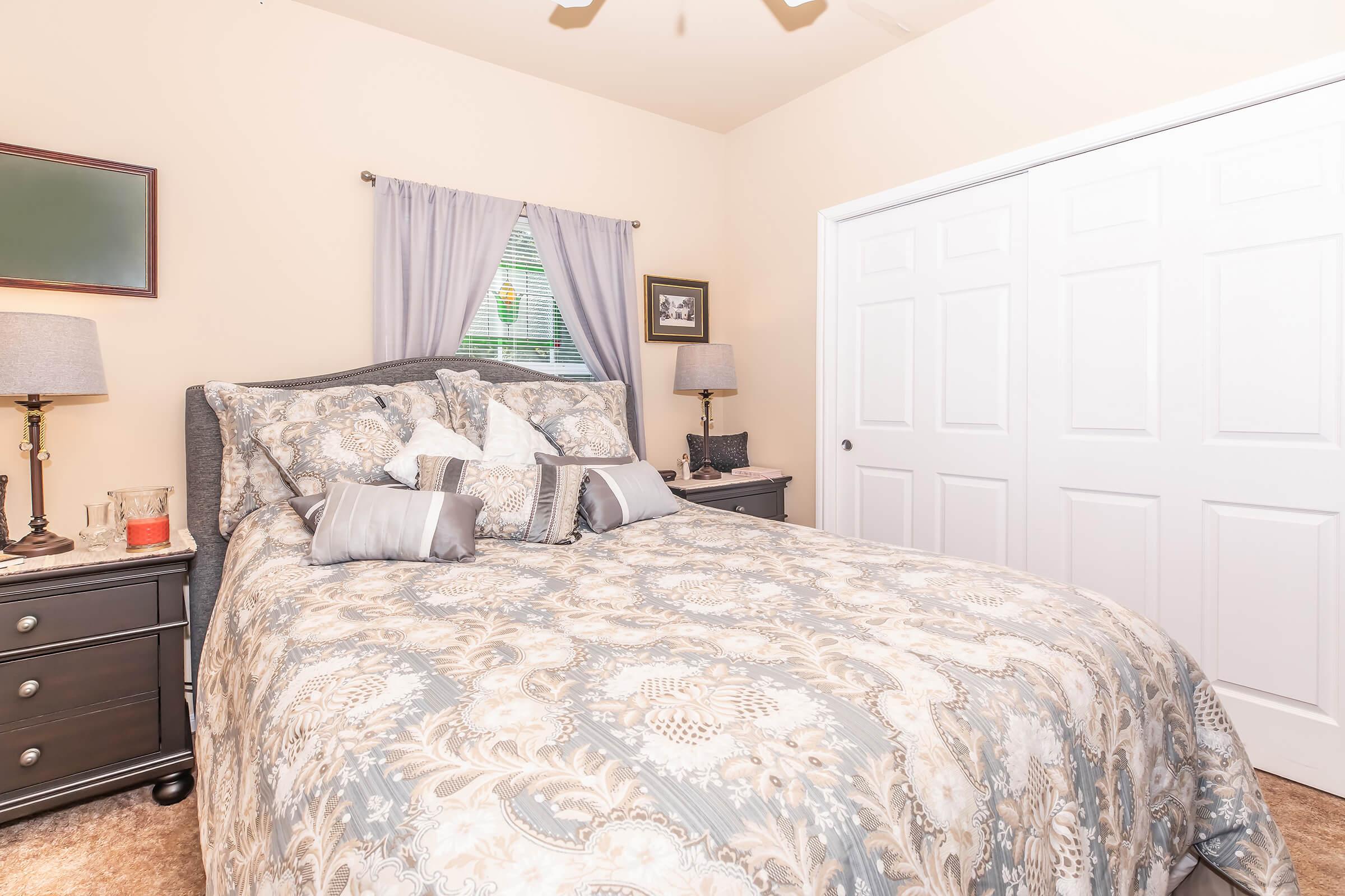 A cozy bedroom featuring a queen-sized bed with a patterned comforter and decorative pillows. Two nightstands with lamps are on either side of the bed, and there's a window with light curtains. A closet with white doors is visible in the background, along with a framed picture on the wall.