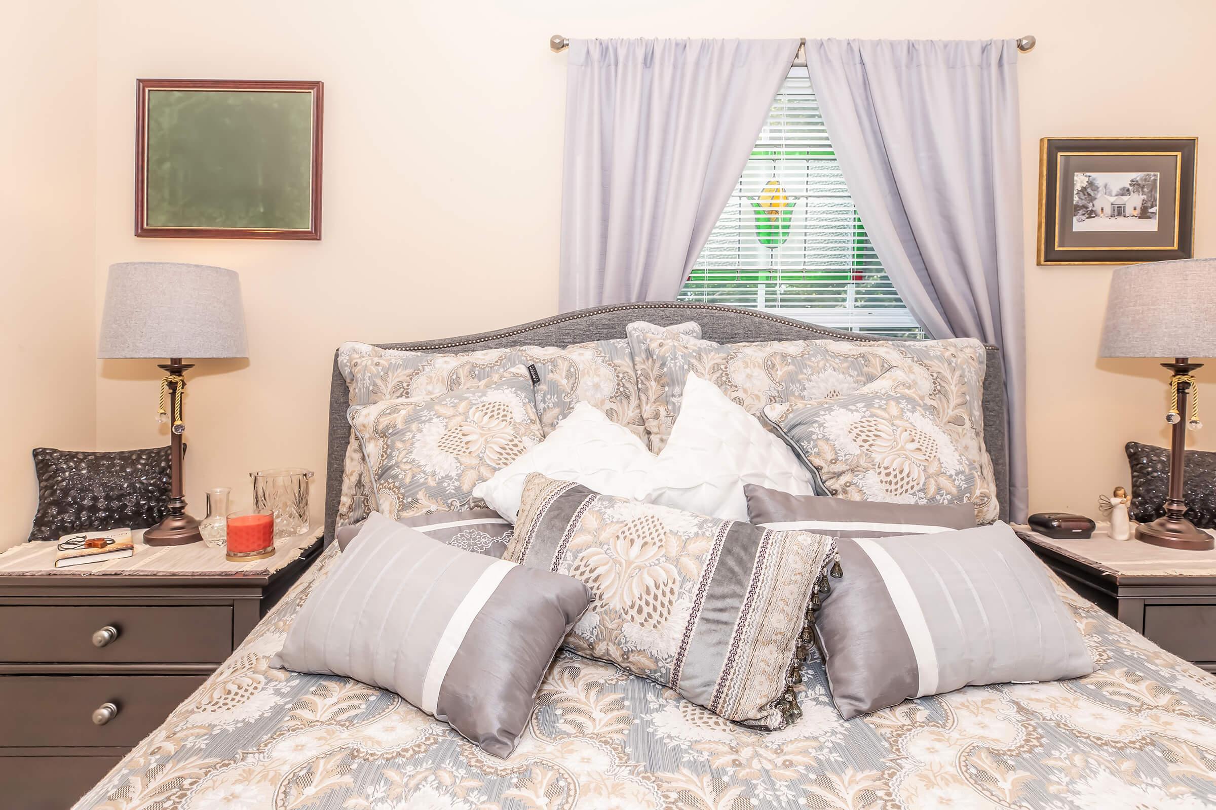 A neatly arranged bedroom featuring a bed with decorative pillows and a floral-patterned comforter. On either side of the bed are nightstands with lamps and various decorative items. A window with soft curtains allows natural light, and framed pictures adorn the walls, creating a cozy atmosphere.