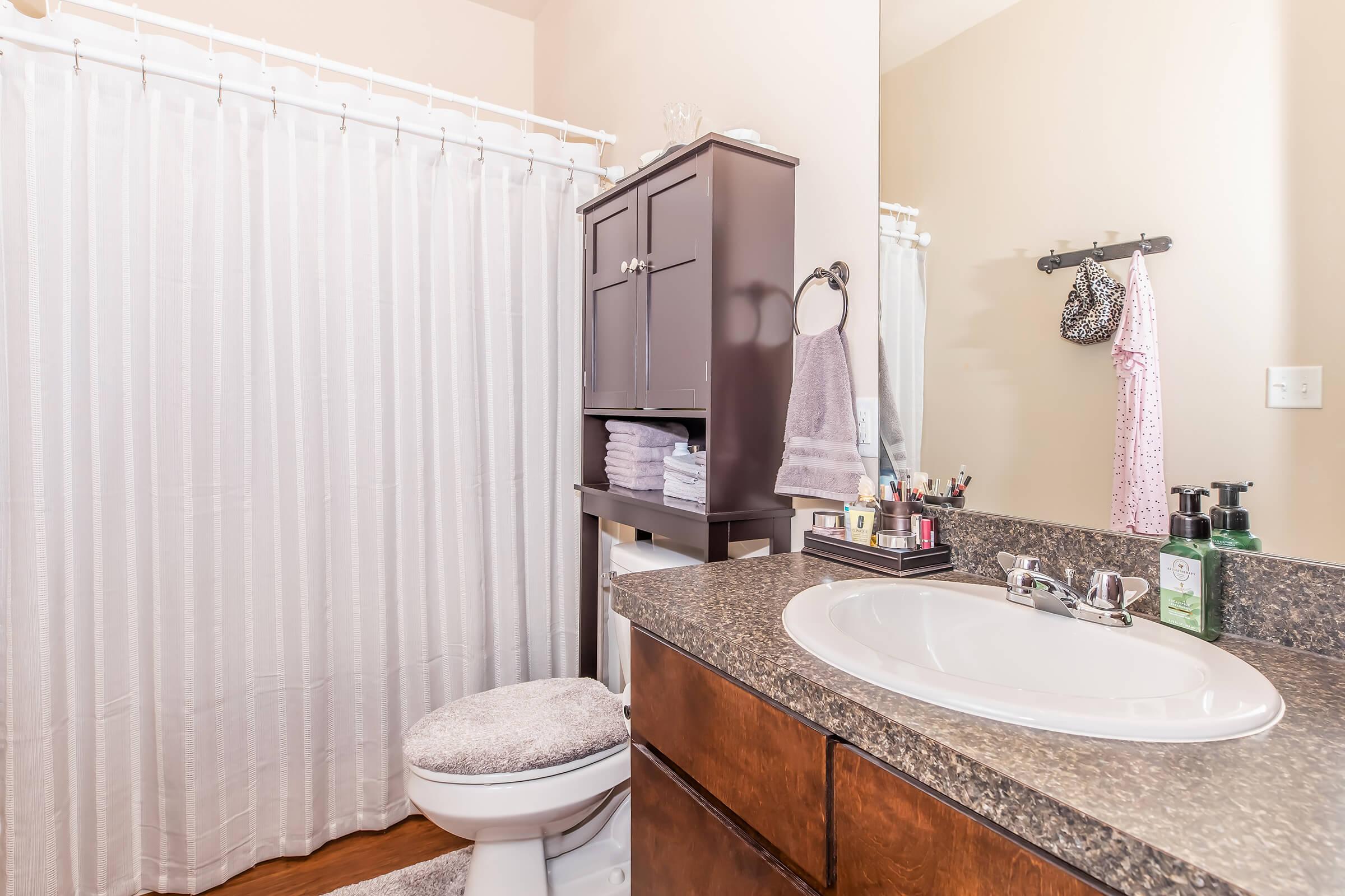 A clean and organized bathroom featuring a white shower curtain, a wooden countertop with a sink, and dark wooden cabinetry. The mirror reflects the space, showcasing neatly stacked towels and various toiletries, creating a serene and functional environment.