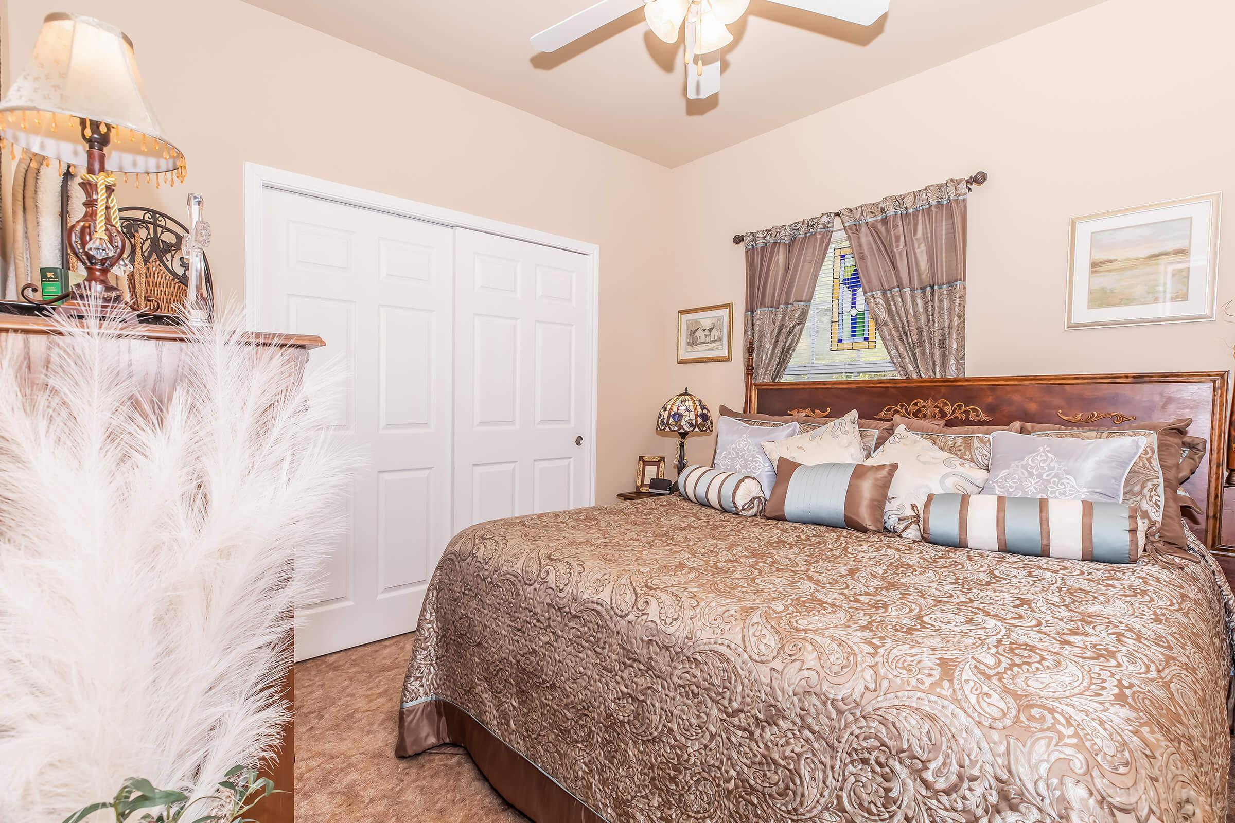 A cozy bedroom featuring a large bed with a decorative brown and gold bedspread and an assortment of patterned pillows. There is a wooden dresser, a lamp, and a ceiling fan. Light-colored walls and a window with curtains complete the warm and inviting atmosphere.