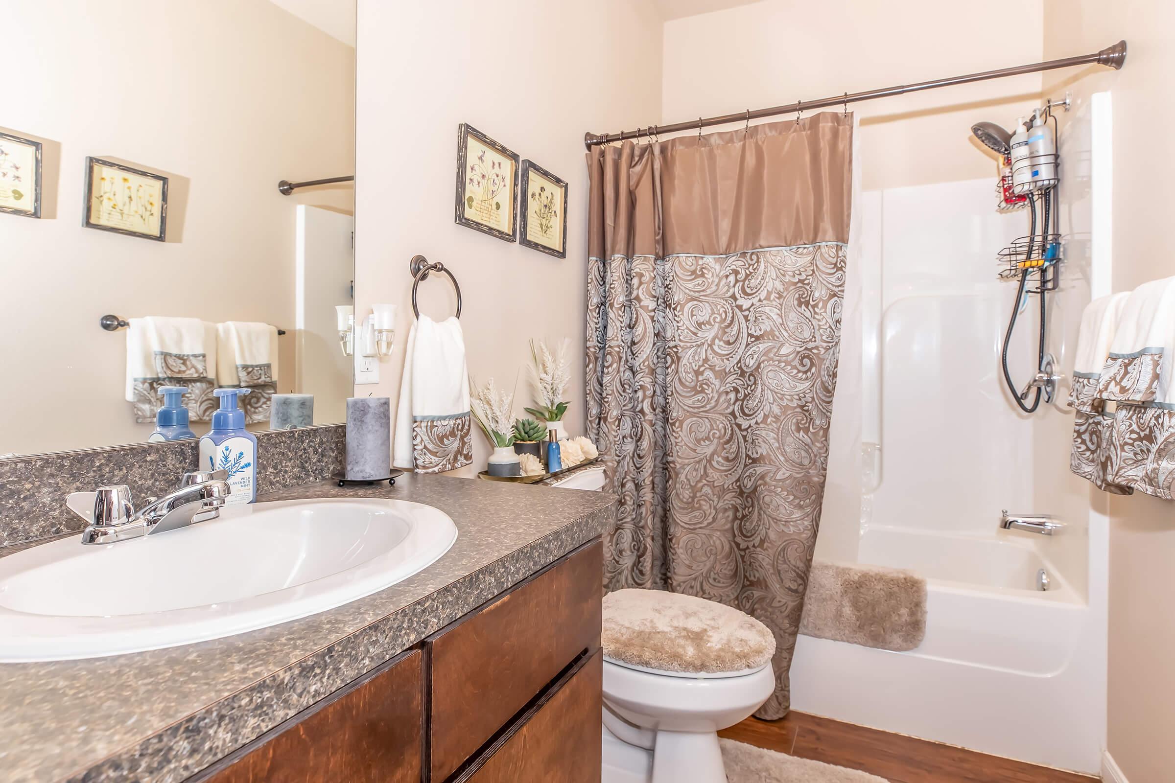 A well-lit bathroom featuring a dark wood vanity with a sink, a circular mirror above it, and decorative toiletries. There is a shower with a patterned curtain, a white bathtub, and plush towels hanging nearby. The walls are painted light beige, enhancing the bright and airy feel of the space.