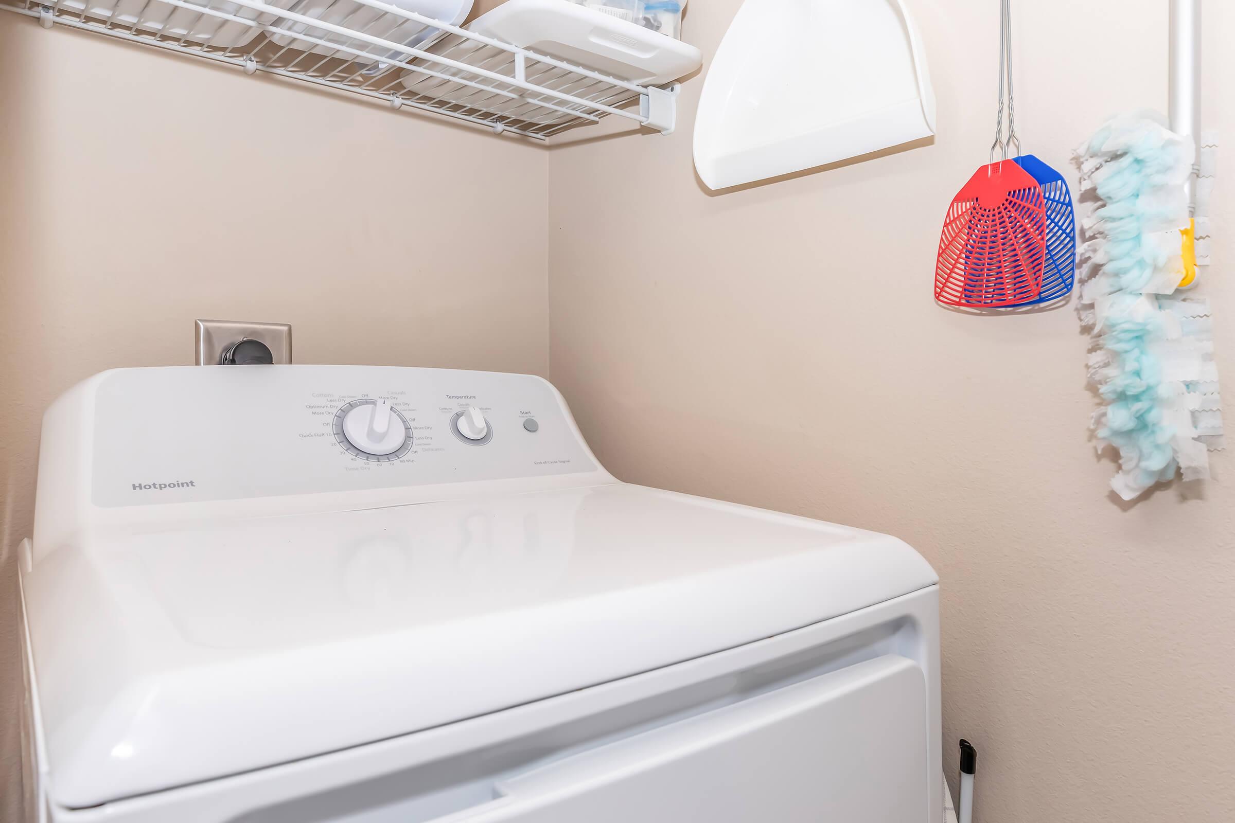 A white washing machine with control knobs is positioned against a beige wall. Above it, there is a wire shelf holding various items, and to the right, a colorful hanging item. There is also a small white scoop attached to the wall, creating a tidy laundry area.