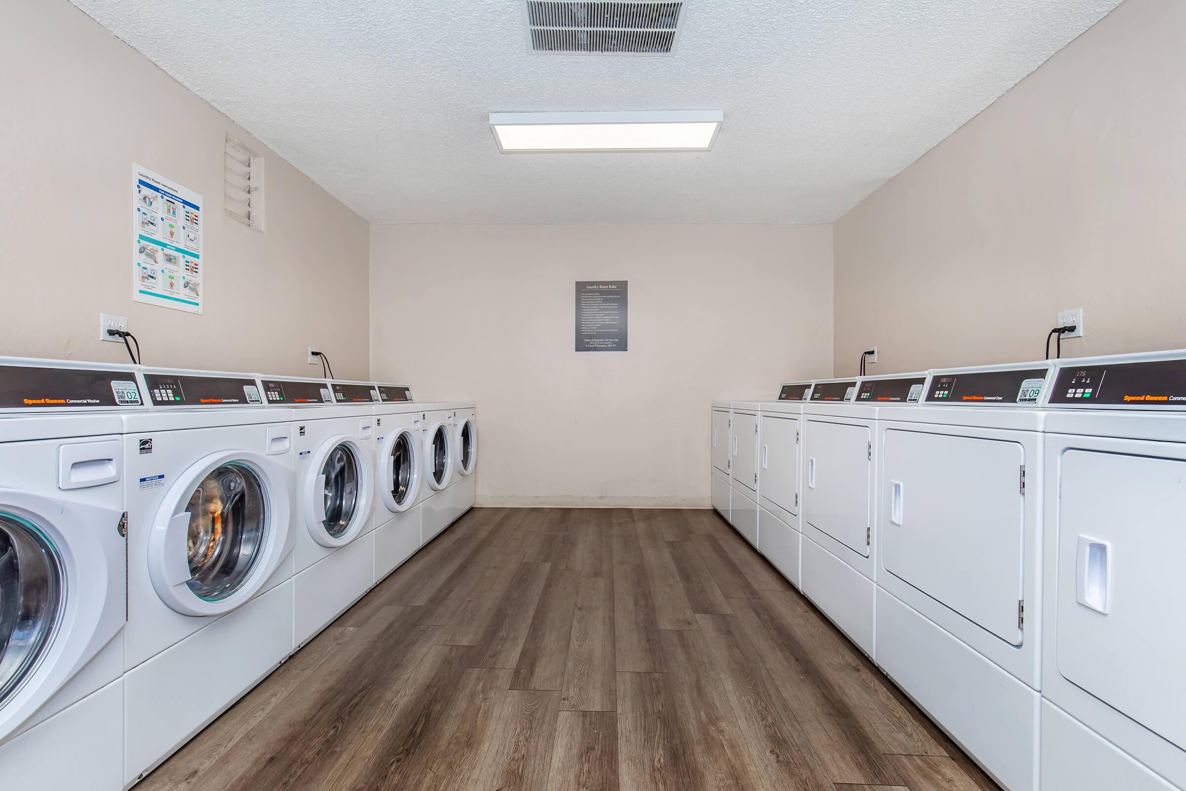 A clean, bright laundry room featuring several rows of white washing machines and dryers along the walls. The flooring is wooden, and there is a soft overhead light. Wall displays provide laundry instructions, and a sign is visible in the center of the room.