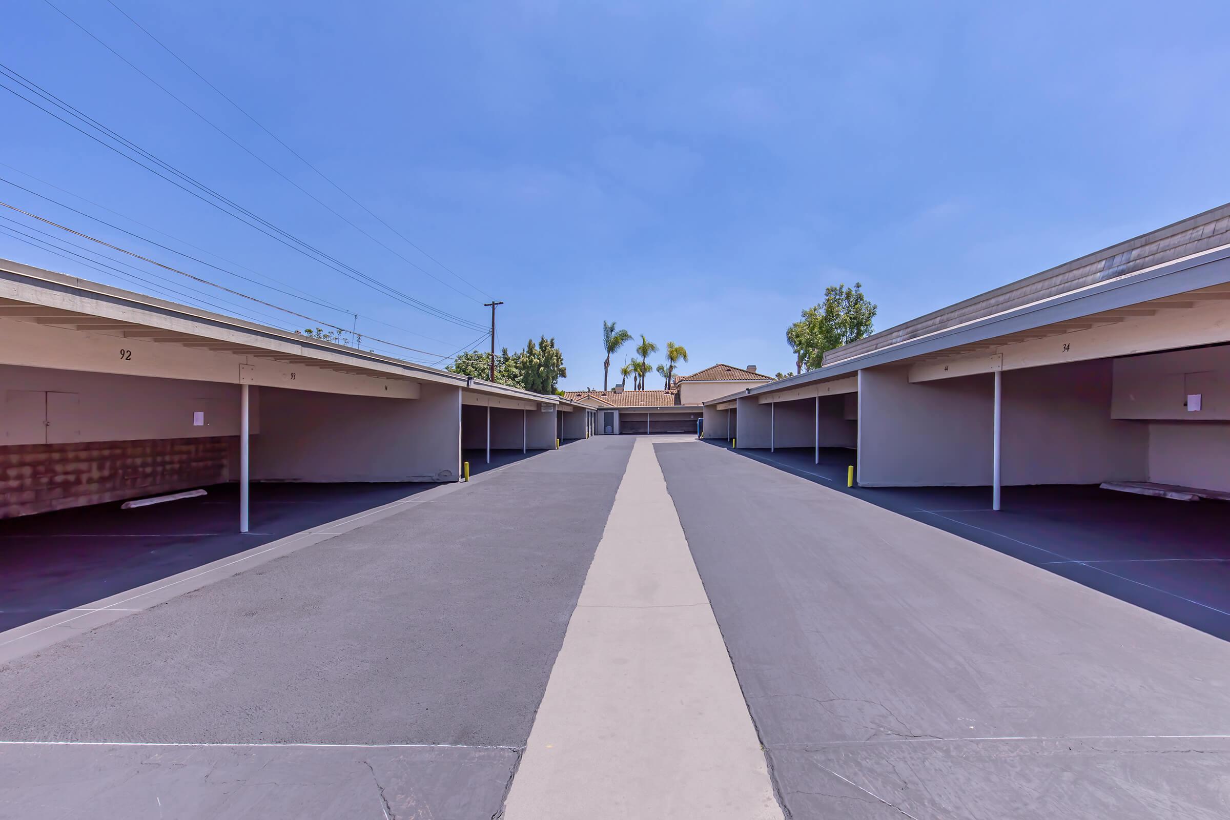 A view of a parking lot featuring several covered parking spaces on either side, with a wide pathway running down the middle. The sky is clear and blue, and there are a few trees and buildings visible in the background. The lot appears orderly and well-maintained.