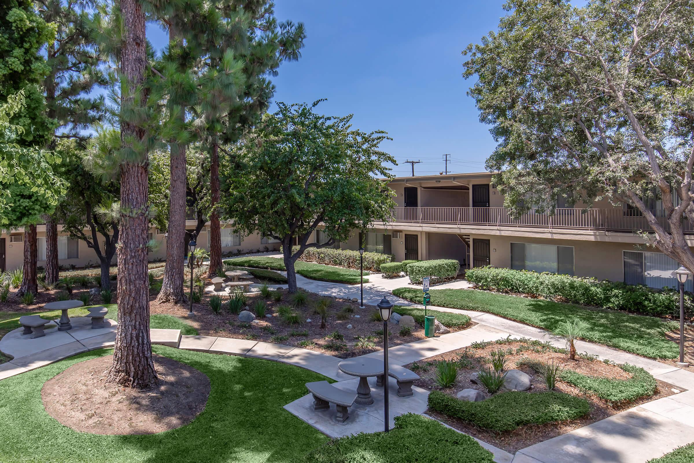 A landscaped courtyard featuring green lawns, trees, and shrubs, surrounded by two-story buildings. There are benches and picnic tables arranged along pathways, providing a serene outdoor space ideal for relaxation and socializing. The sky is clear and blue, enhancing the peaceful atmosphere.