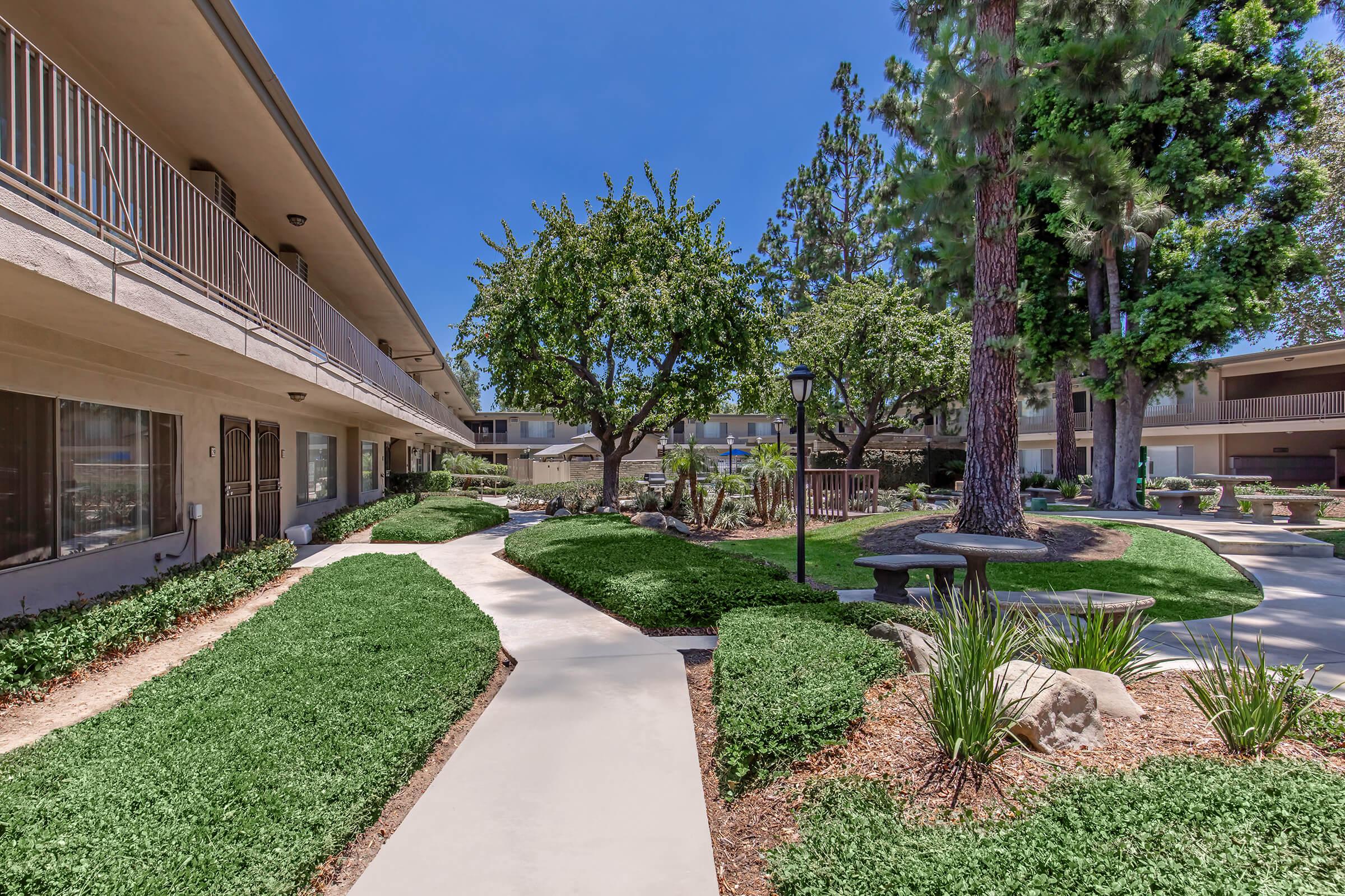 A well-maintained outdoor area of an apartment complex featuring a concrete pathway winding through manicured lawns, shrubs, and trees. Surrounding balconies of the building overlook the landscaped garden, with a small table and chairs set under a tree for relaxation. The sky is clear and blue.