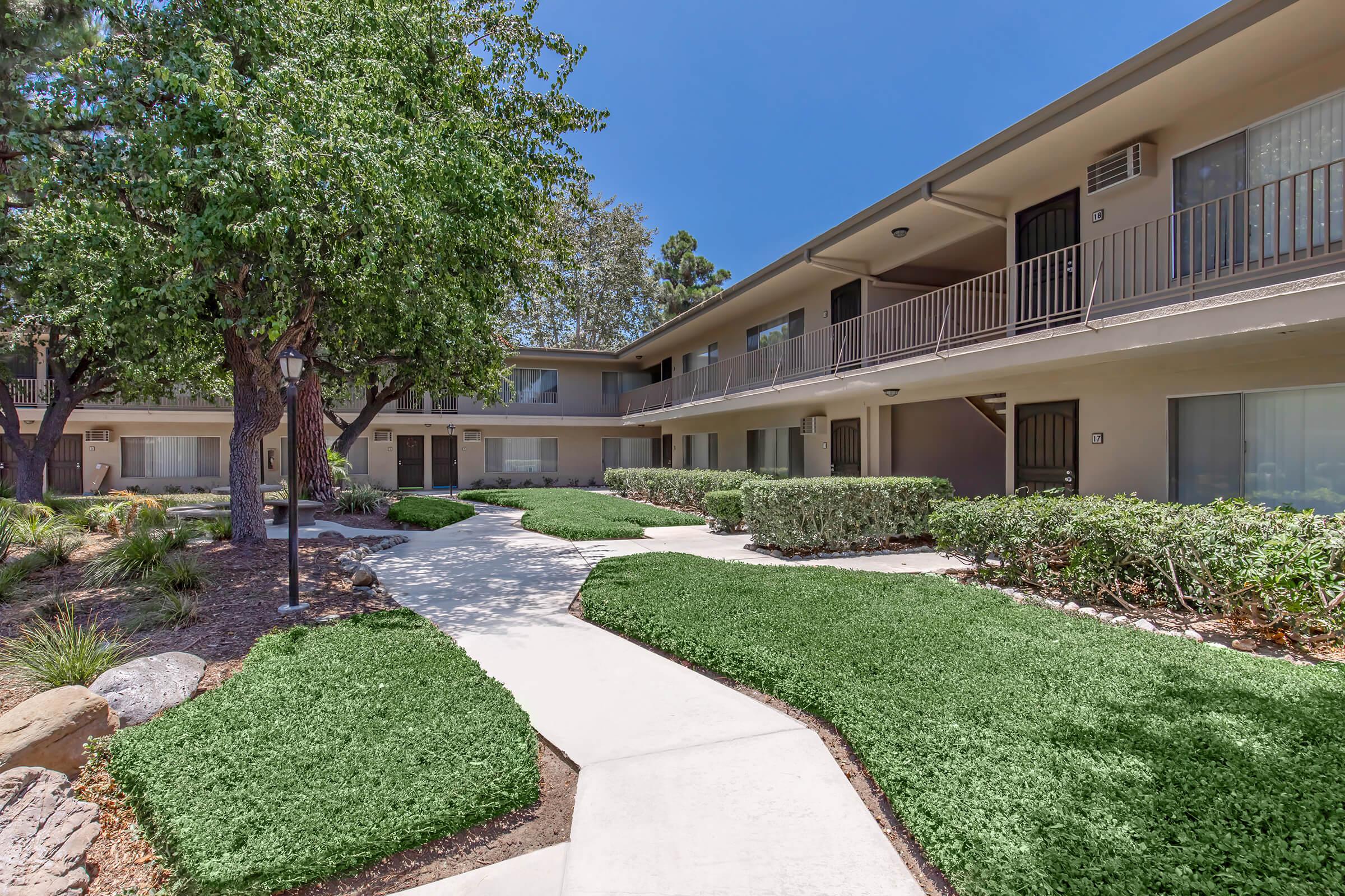A well-maintained outdoor area featuring a curved walkway surrounded by green grass and shrubs, leading to two-story apartment buildings. The scene is bright with clear blue skies and plenty of trees providing shade. The overall atmosphere is inviting and peaceful.