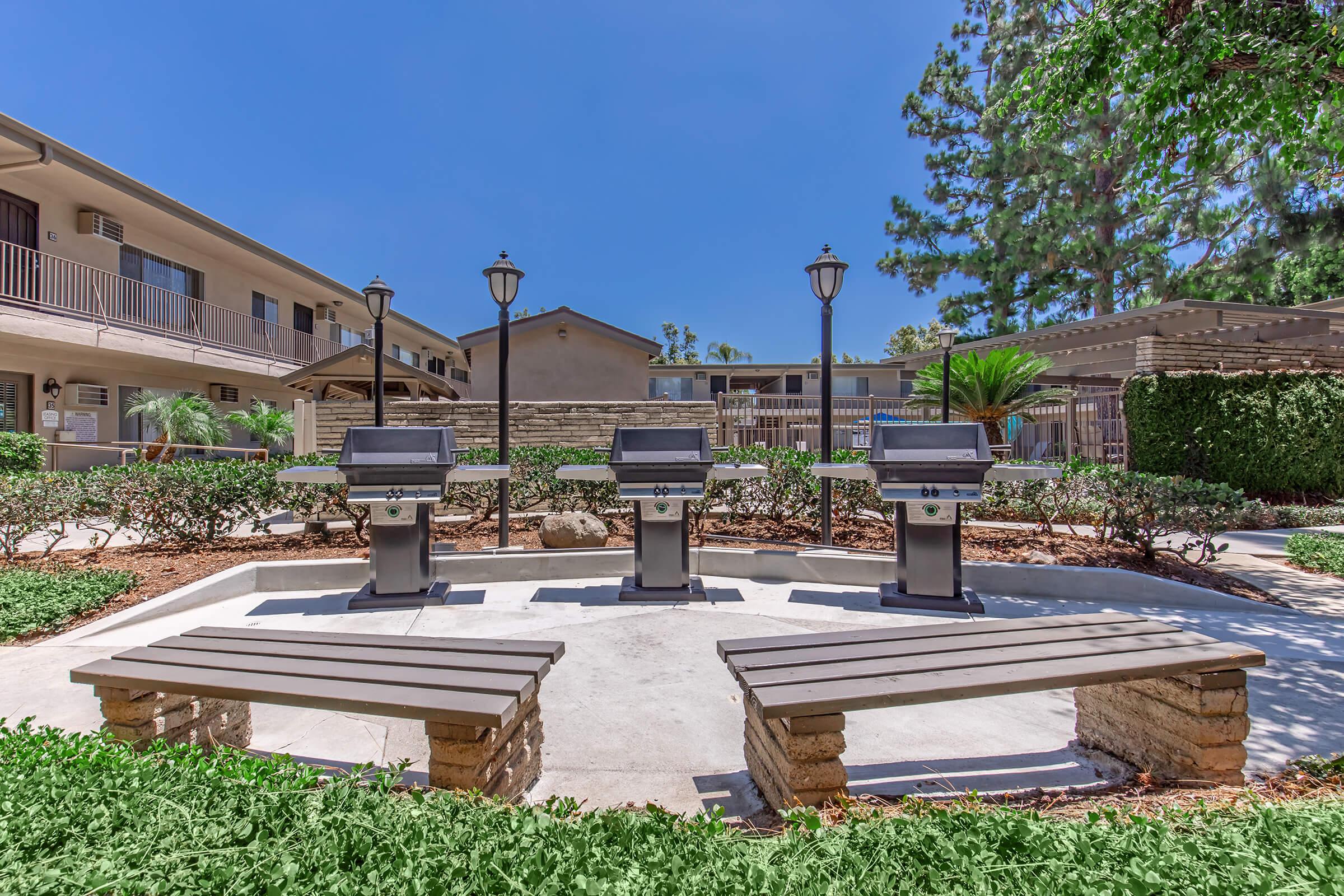 Grilling area featuring four stainless steel barbecue grills surrounding a circular concrete space with benches. Lush greenery and decorative shrubs are present, along with several lampposts. In the background, there are multi-story buildings under a clear blue sky.
