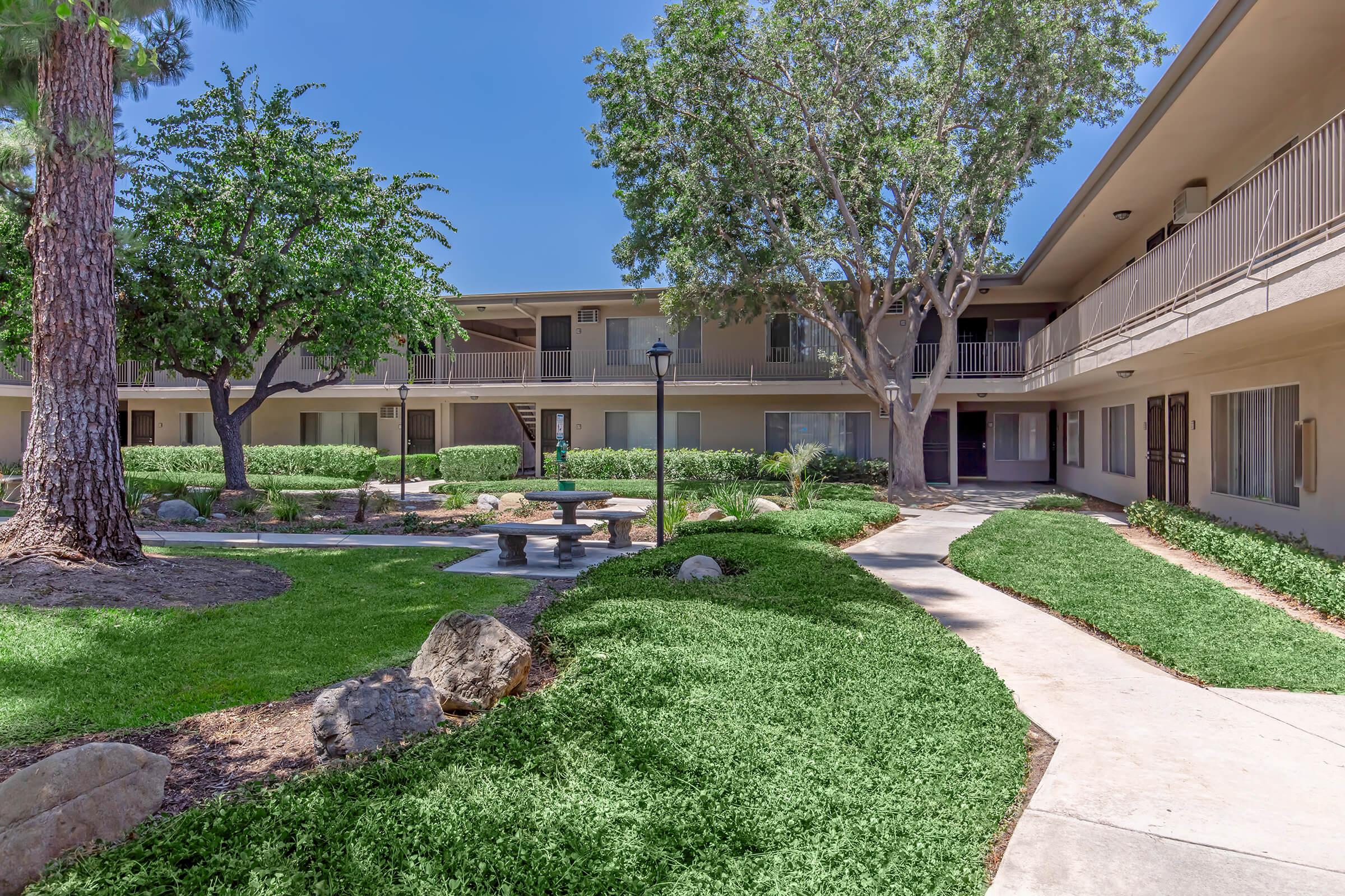 A sunny courtyard of an apartment complex featuring landscaped greenery, small trees, and a stone bench surrounded by pathways. The building is two stories high, with a clear blue sky above and walkways connecting the upper and lower levels.