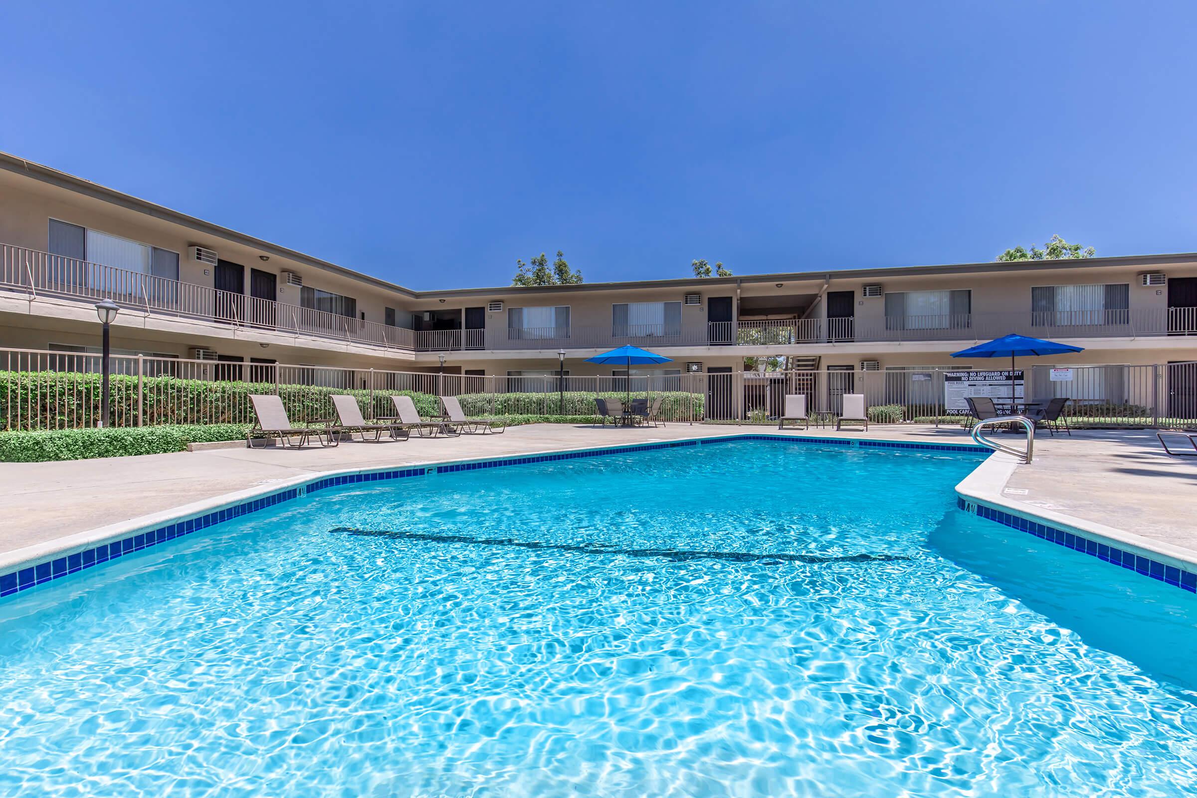 A vibrant swimming pool surrounded by lounge chairs, with a two-story building in the background featuring balconies. Bright blue skies and landscaped greenery enhance the relaxing atmosphere of the pool area.