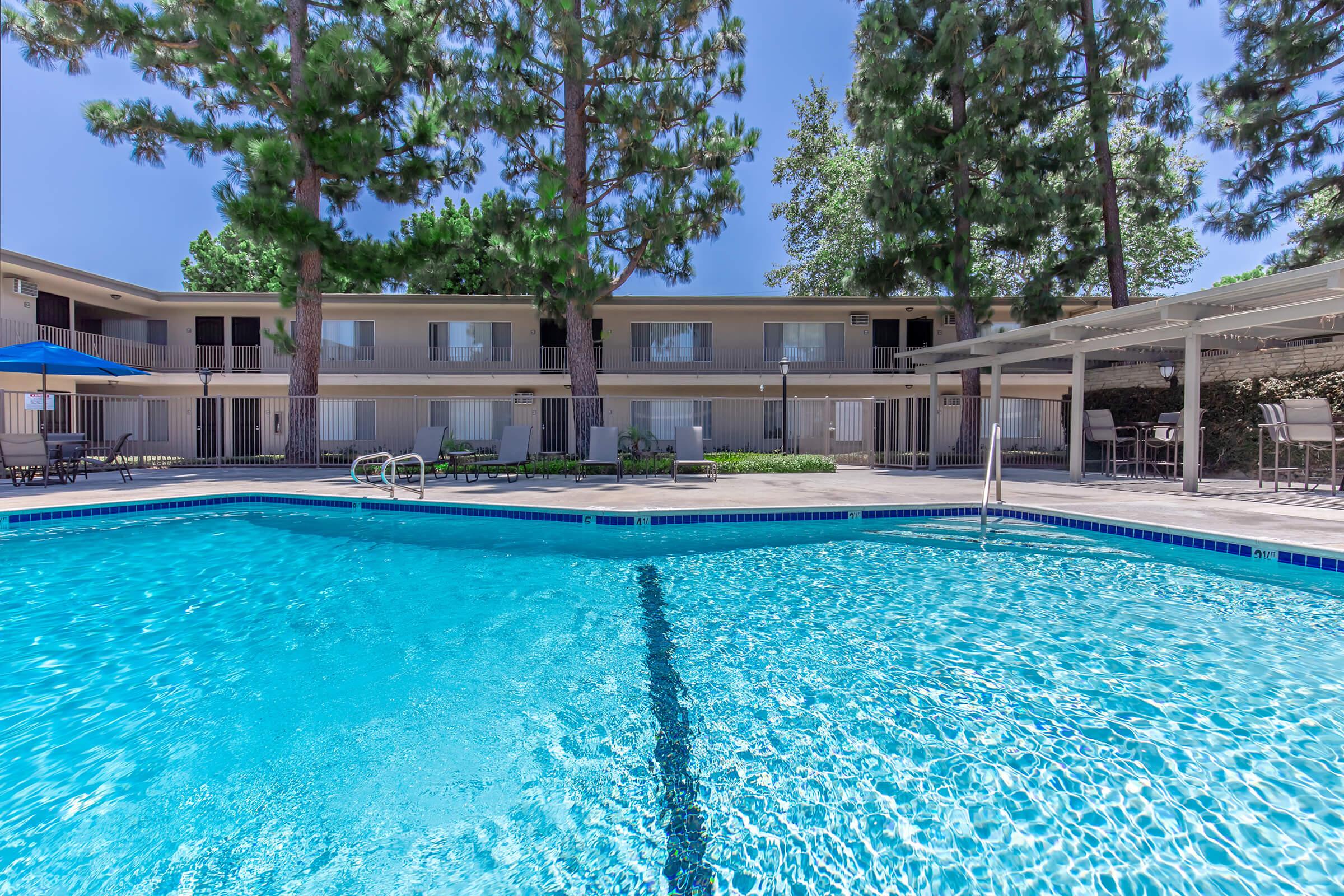 A clear blue swimming pool surrounded by lounge chairs, with tall green trees in the background. Two-story buildings are visible, featuring balconies and large windows. An umbrella provides shade near the pool area, creating a relaxing outdoor atmosphere.
