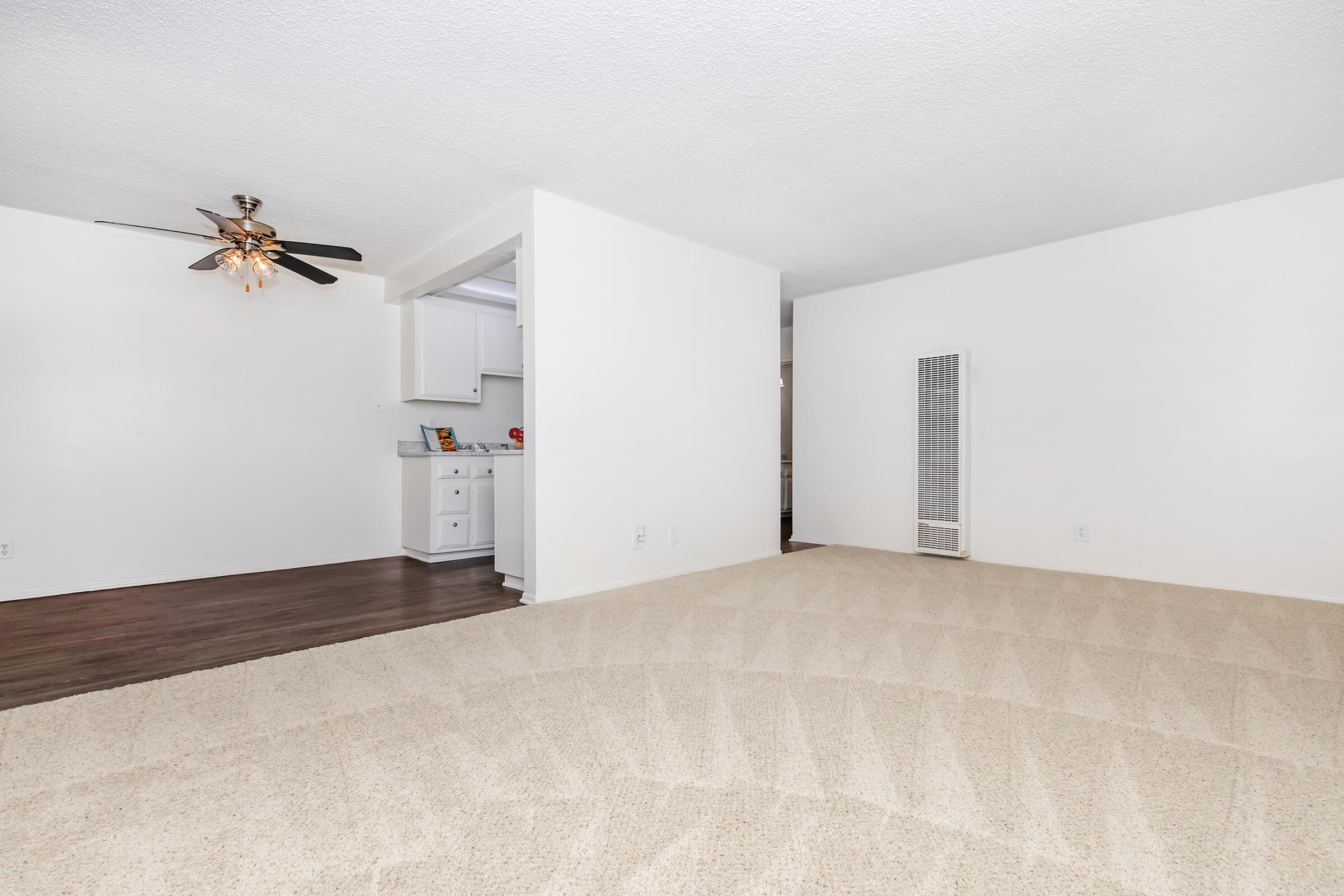 Spacious living area with light beige carpet, featuring a ceiling fan, adjacent to a kitchen area visible through an open entrance. The walls are painted white, and there is a vertical radiator against the far wall. Natural light creates a bright, airy atmosphere in the room.